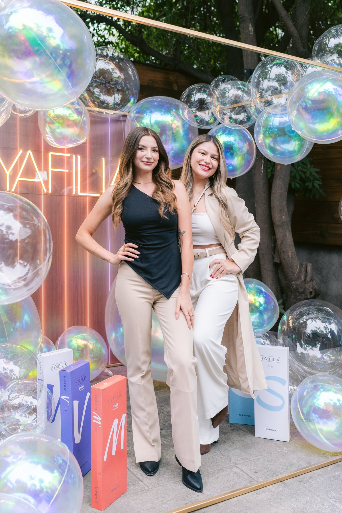 Dos mujeres posando en una celebración con globos transparentes y un fondo de luces y una pared de madera. Una de ellas lleva pantalones beige y blusa negra, la otra pantalones blancos y chaqueta beige.