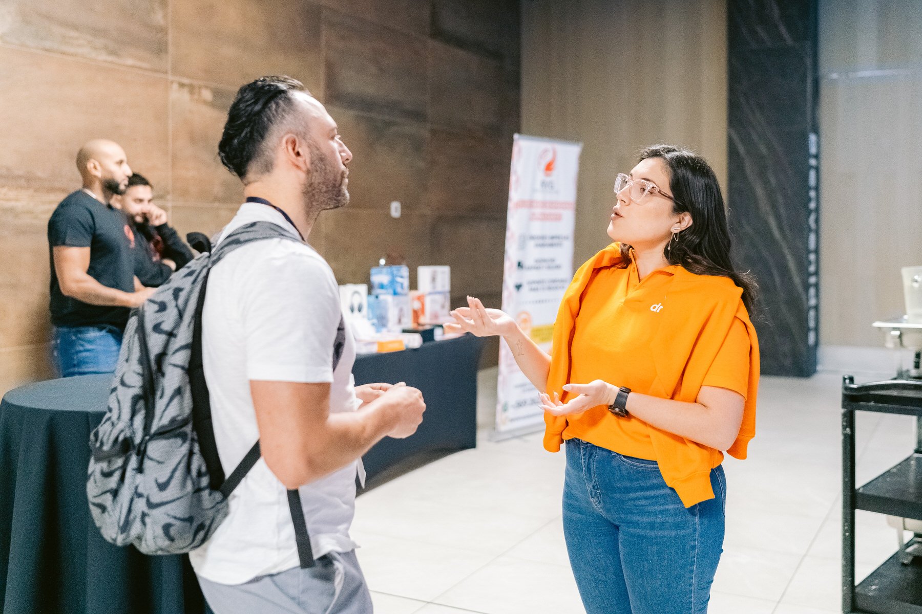 Una mujer de cabello oscuro, gafas y camiseta naranja, habla con un hombre con mochila en un evento en interiores, con una mesa y un banner en el fondo.