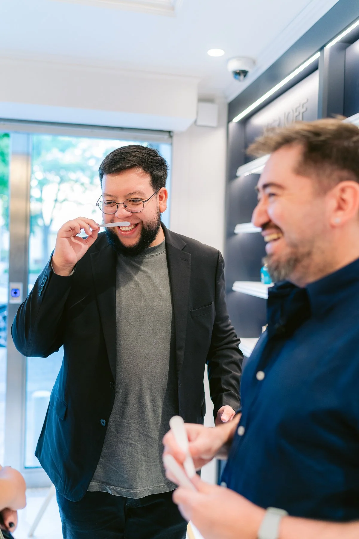 Dos hombres riendo y conversando en lugar moderno, uno con gafas, el otro usando una camisa azul, uno de ellos con un bolígrafo.