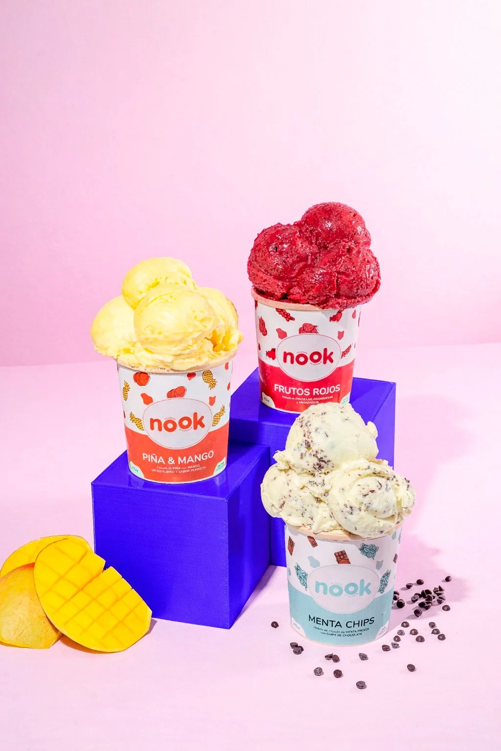 Tres copas de helado de diferentes sabores y colores, colocadas sobre una estructura azul. La copa de la izquierda tiene helado amarillo con sabor a piña y mango, la del centro tiene helado rojo de frutos rojos, y la derecha tiene helado blanco con c