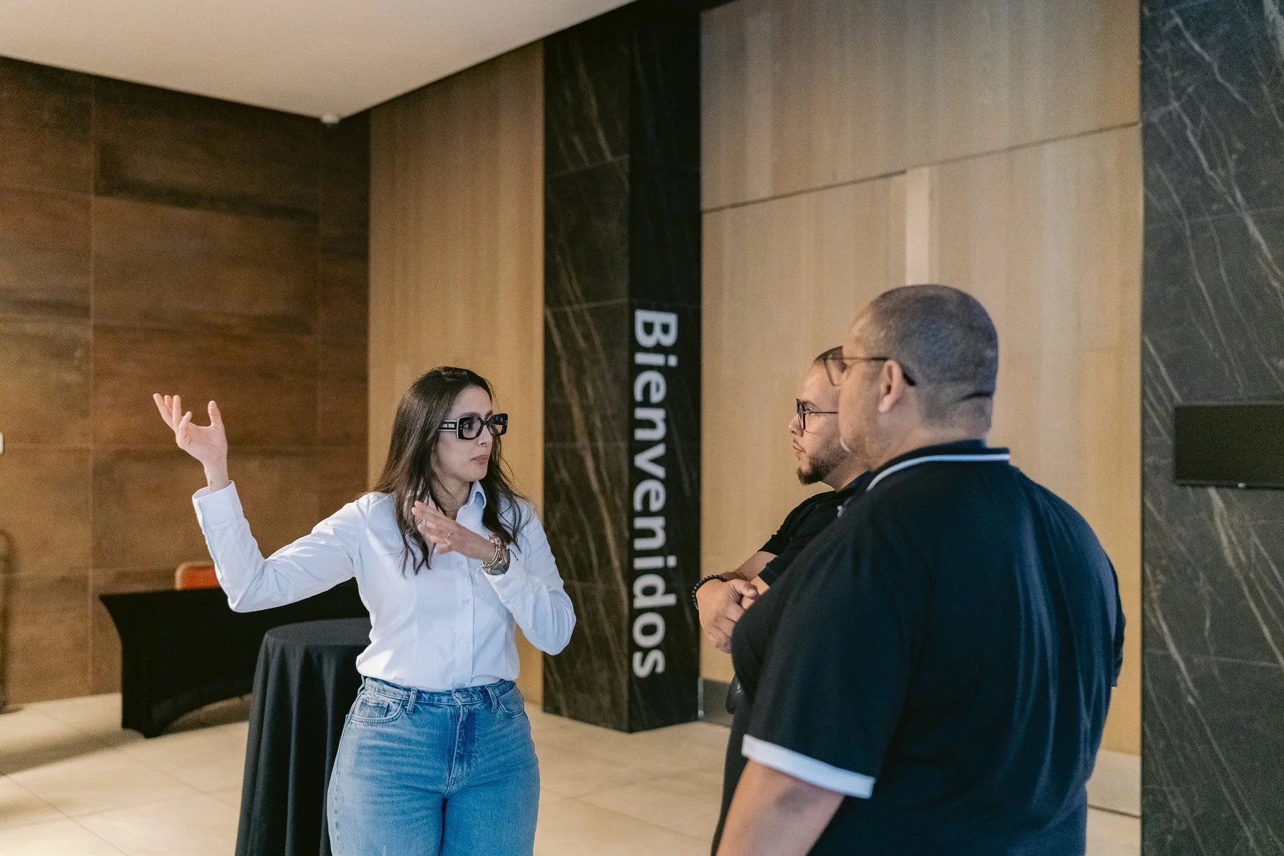 Tres personas hablando en una conferencia o reunión, una mujer explicando y dos hombres escuchando, en un lugar con cartel que dice 'Bienvenida Sociedad'.
