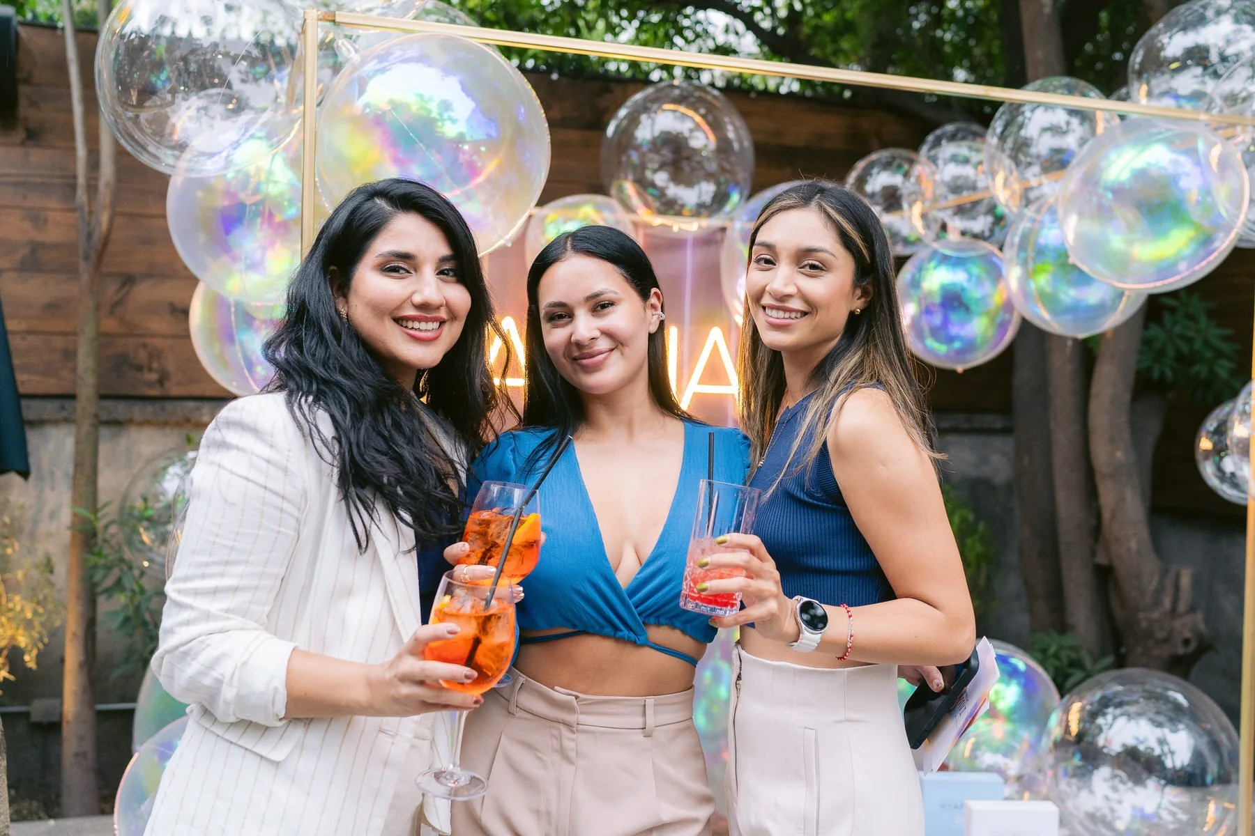 Tres mujeres sonriendo y sosteniendo bebidas en un evento al aire libre con decoraciones de globos y luces