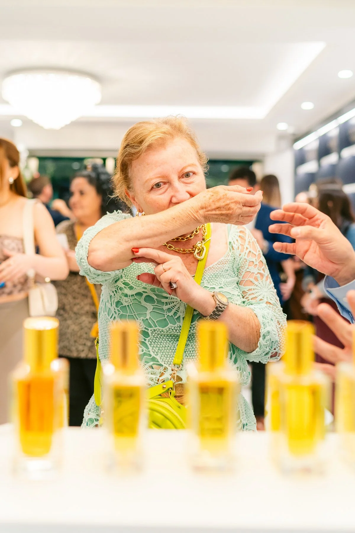 Mujer mayor oliendo una muestra de perfume en una tienda, rodeada de otras personas y frascos de perfume en primer plano.