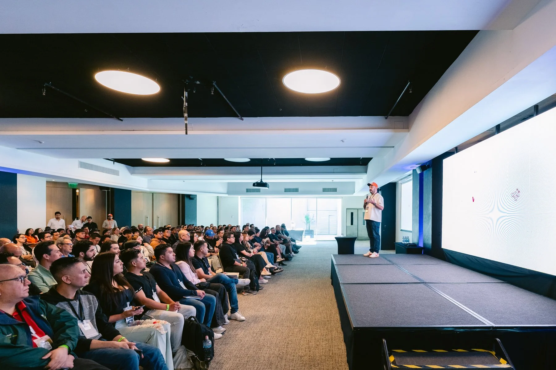 Hombre dando una charla en un escenario frente a una audiencia en una conferencia o seminario en un auditorio con pantalla grande.