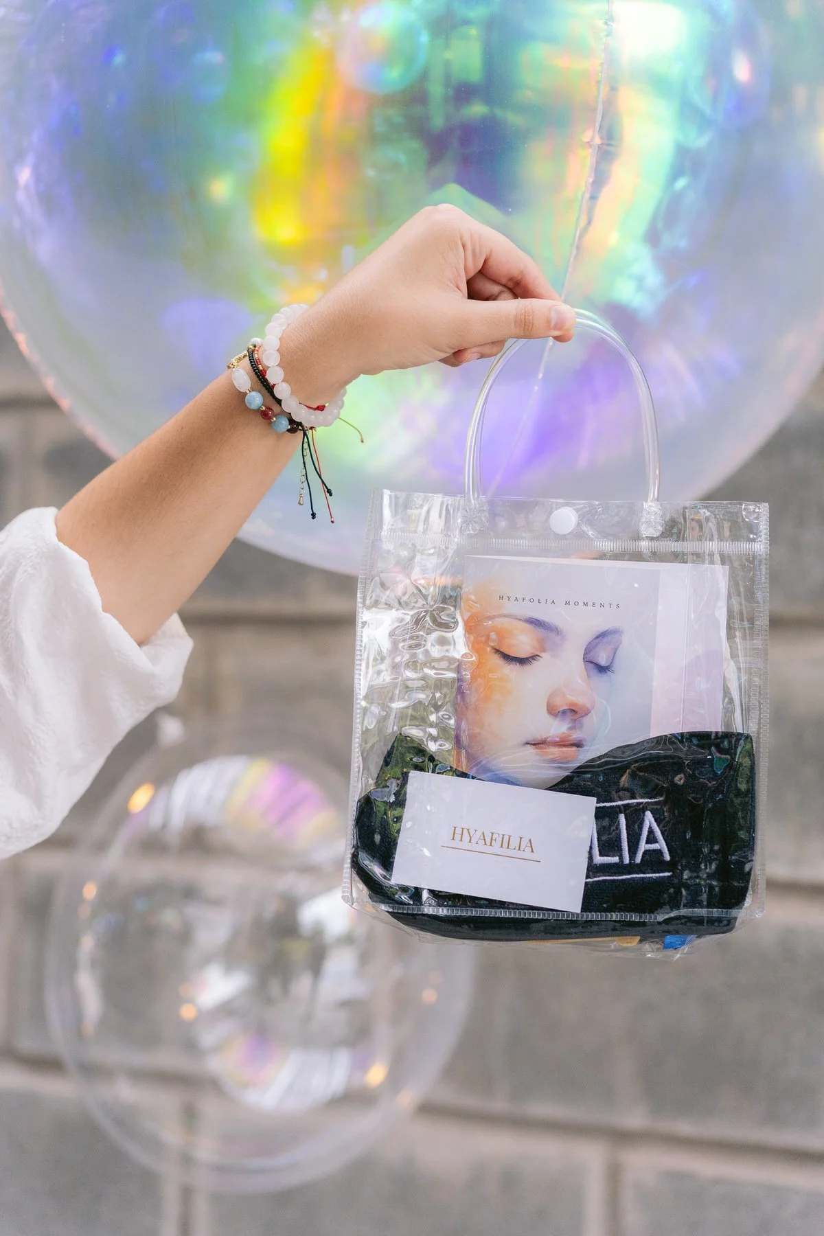 Mano sosteniendo una bolsa de plástico transparente con una caja y una prenda de ropa con la marca HYAFILIA. Fondo con globos de colores iridiscentes.