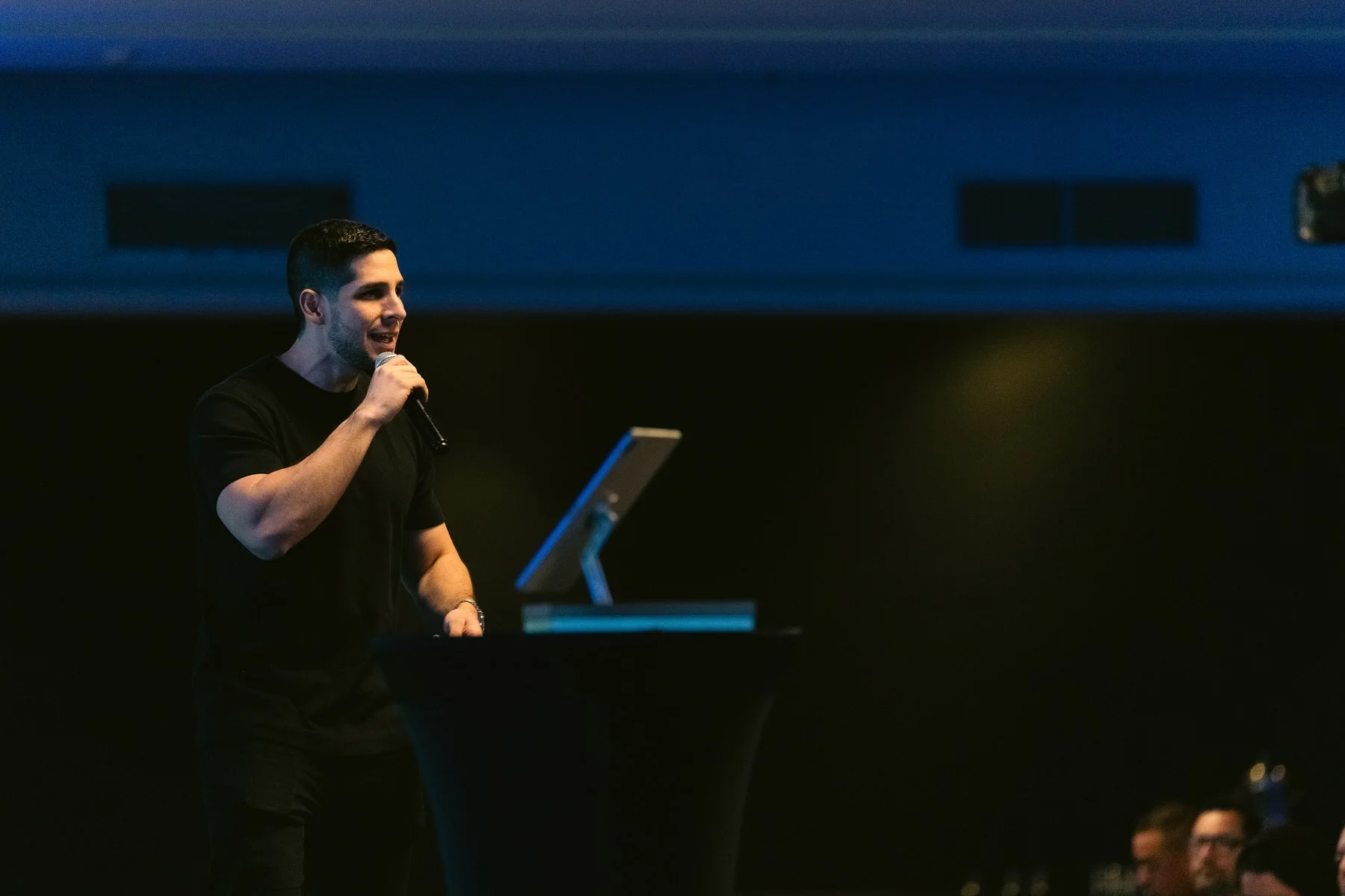 Joven hombre hablando con micrófono en un escenario oscuro.