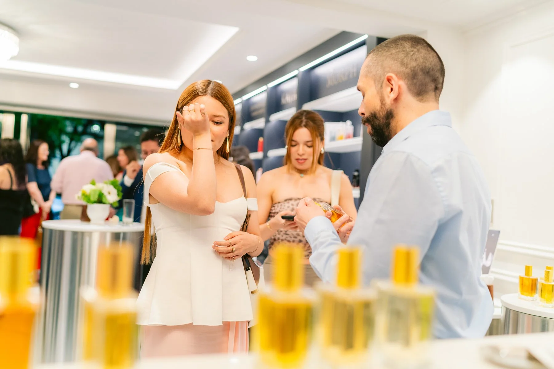 Mujer rubia con vestido blanco probándose perfume en una tienda o exhibición, rodeada de otras personas en el fondo.