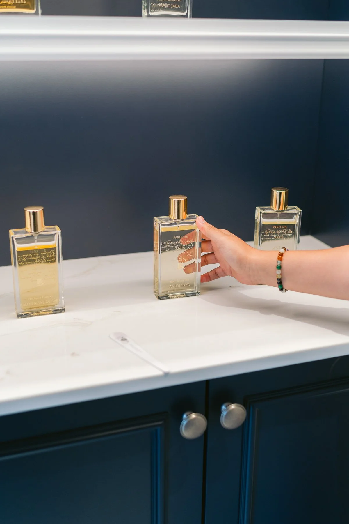 Mano de mujer con pulsera de cuentas, sosteniendo un frasco de perfume en una exhibición con otros frascos similares sobre un mostrador blanco.