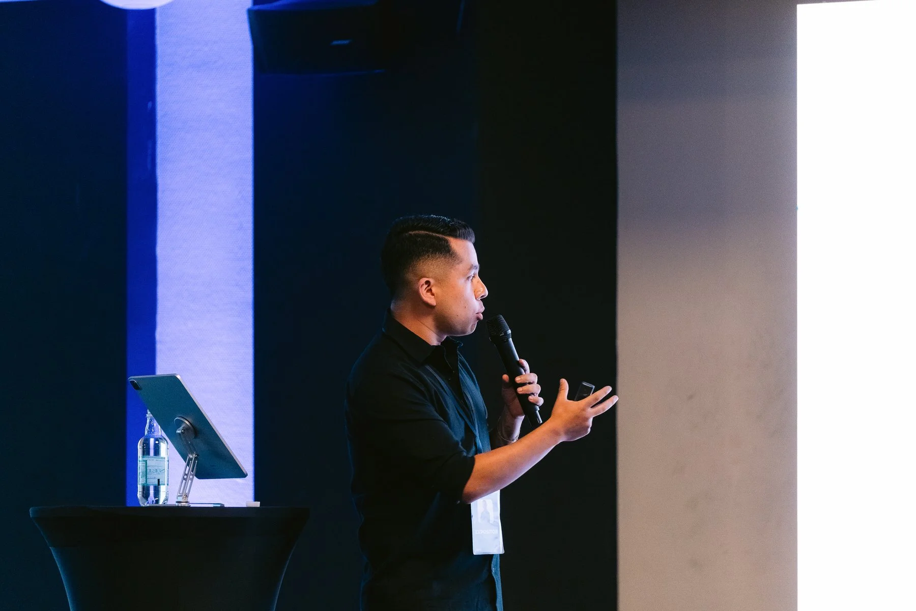 Hombre en escenario presentando una diapositiva, sosteniendo un micrófono y un teléfono, con una botella de agua y un dispositivo en un stand a su lado.
