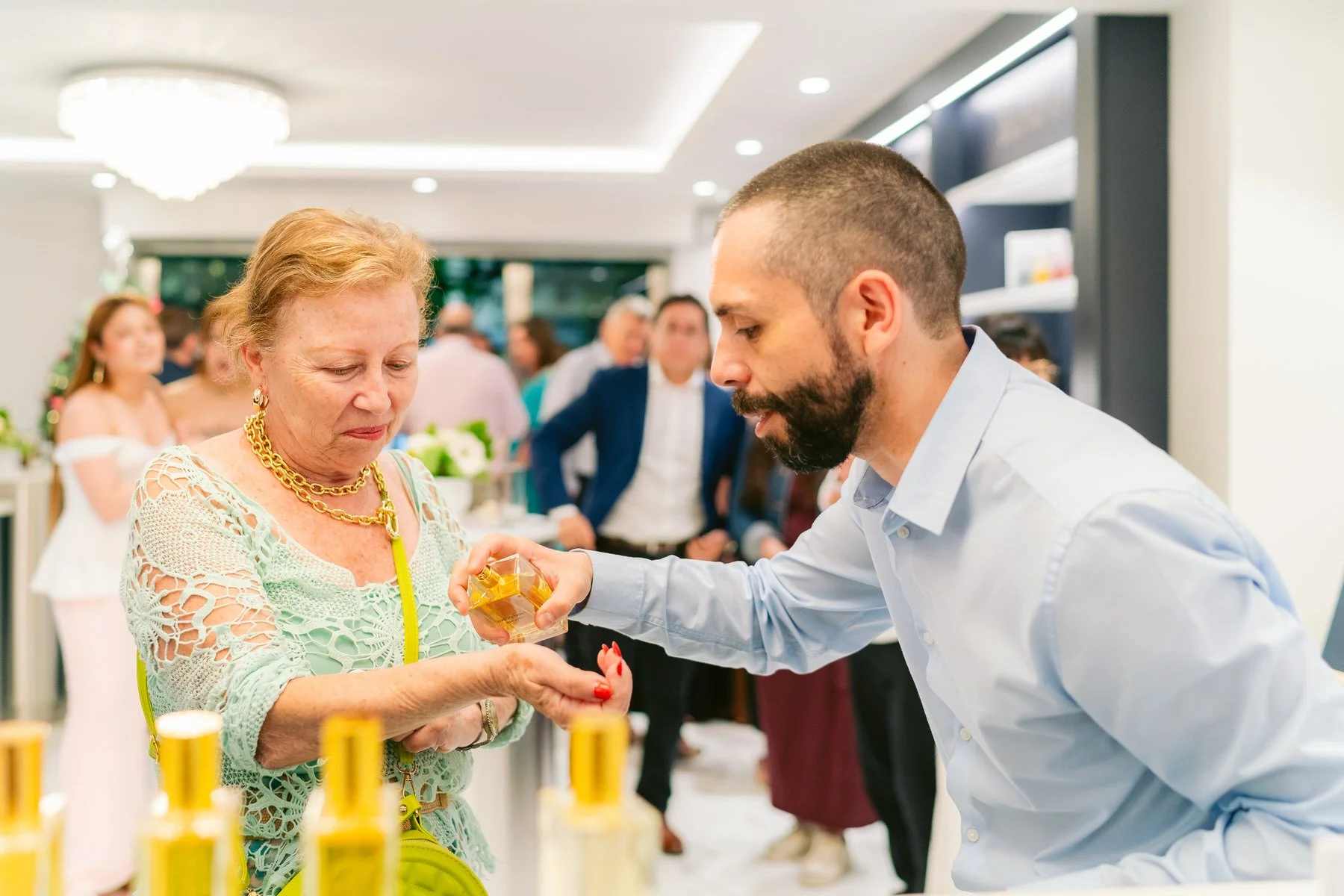 Una mujer mayor recibe muestra de perfume en una tienda o evento, mientras un hombre le ayuda.