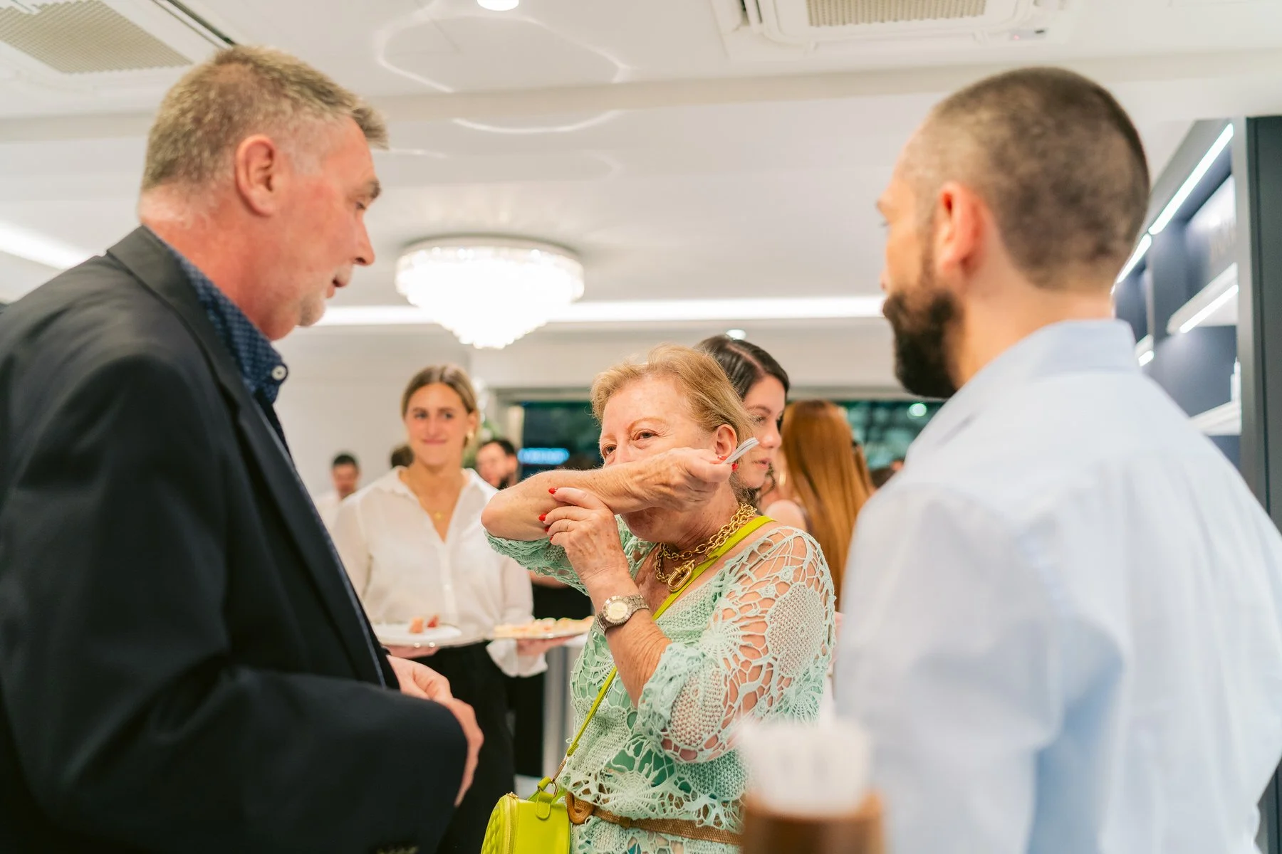 Persona mayor riendo y cubriéndose la boca con la mano durante un evento social, rodeada de varias personas en un ambiente elegante.