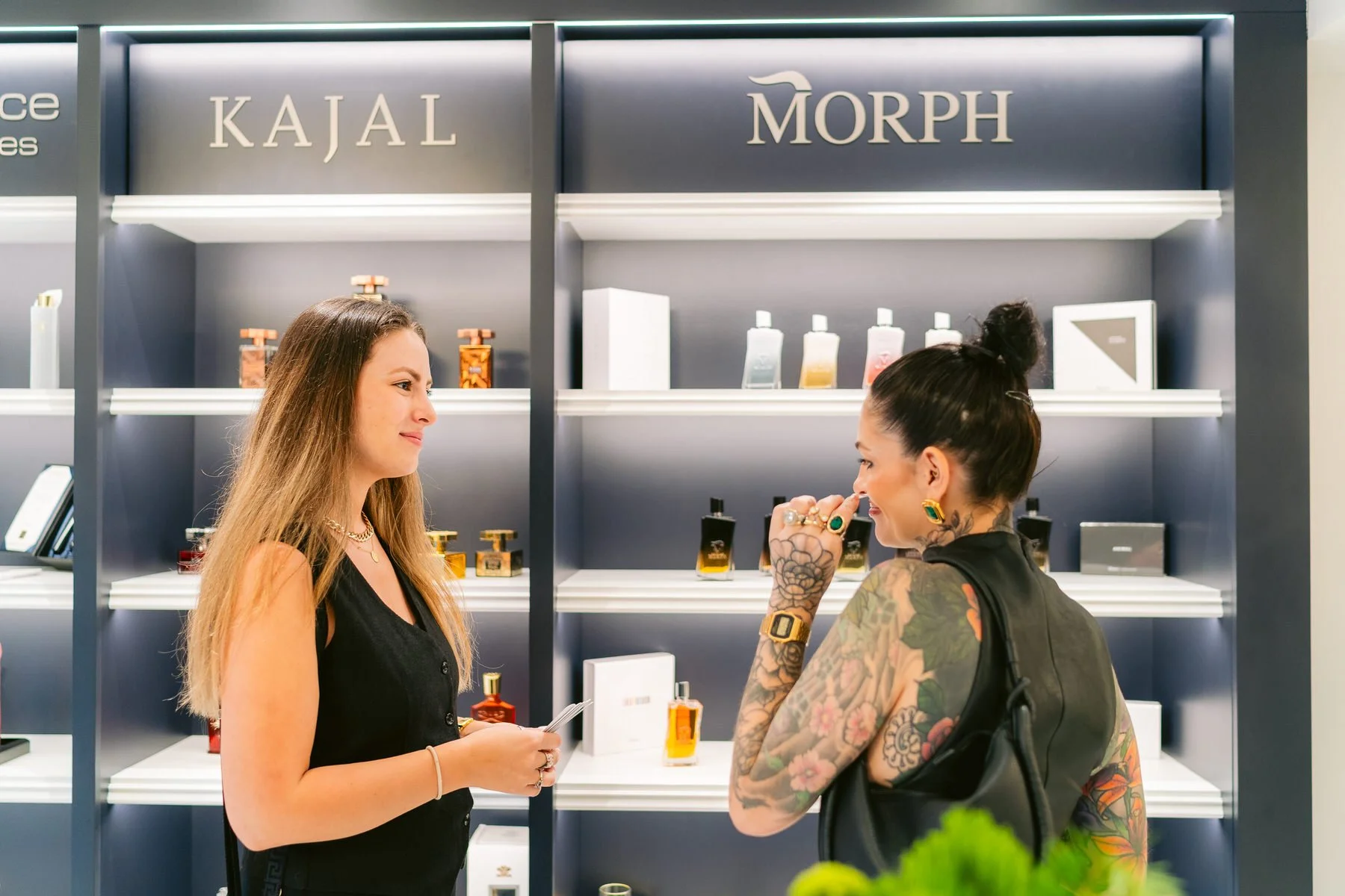 Dos mujeres conversando en una perfumería, una con tatuajes y otra con cabello largo, frente a estantes con botellas de perfume y accesorios.