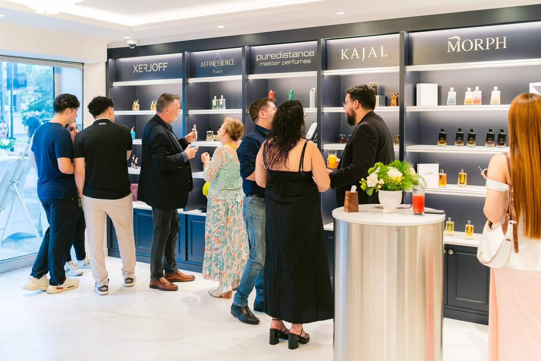 Varias personas en una tienda de perfumes, algunos sosteniendo y olfateando frascos, en un ambiente bien iluminado con estanterías de perfumes y un mostrador con flores y bebidas.