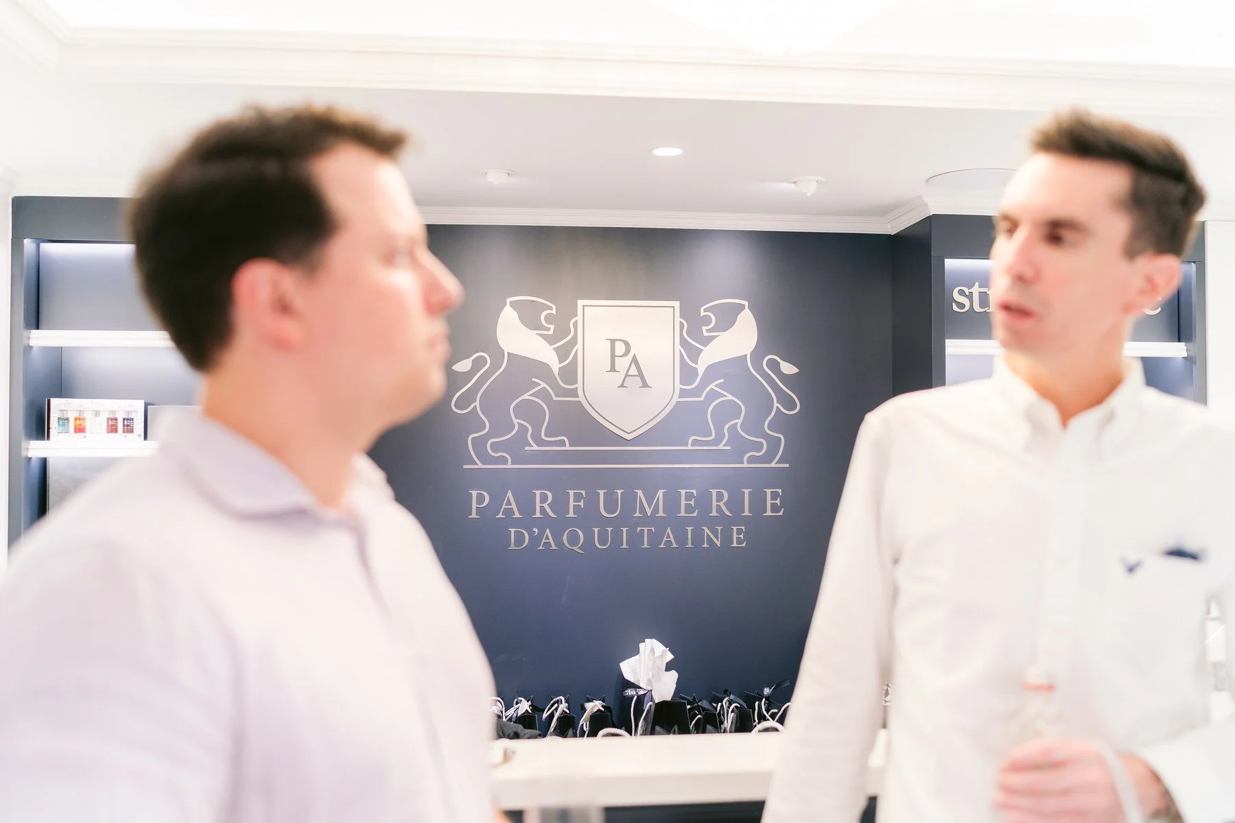 Dos hombres conversando en una perfumería llamada Parfumerie d'Aquitaine, con un fondo que muestra un logotipo con dos leones y letras 'PA'.