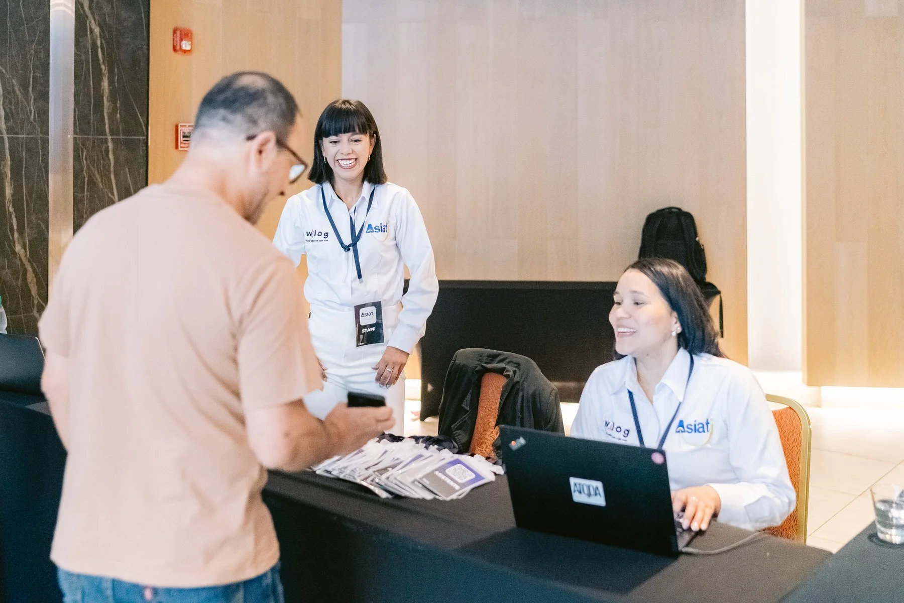 Una mujer sonriente en uniforme de staff hablando con un hombre en una mesa de registro en un evento.