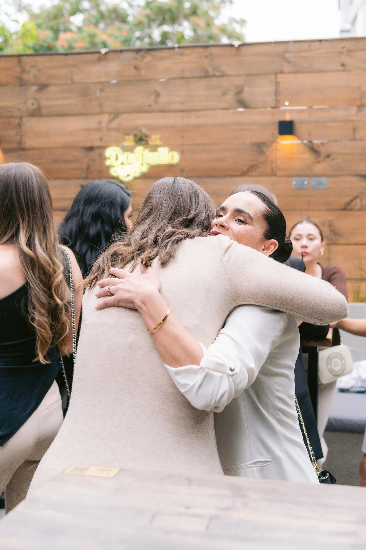 Dos mujeres se abrazan con cariño en un ambiente social, en un lugar con paredes de madera y decoraciones modernas.