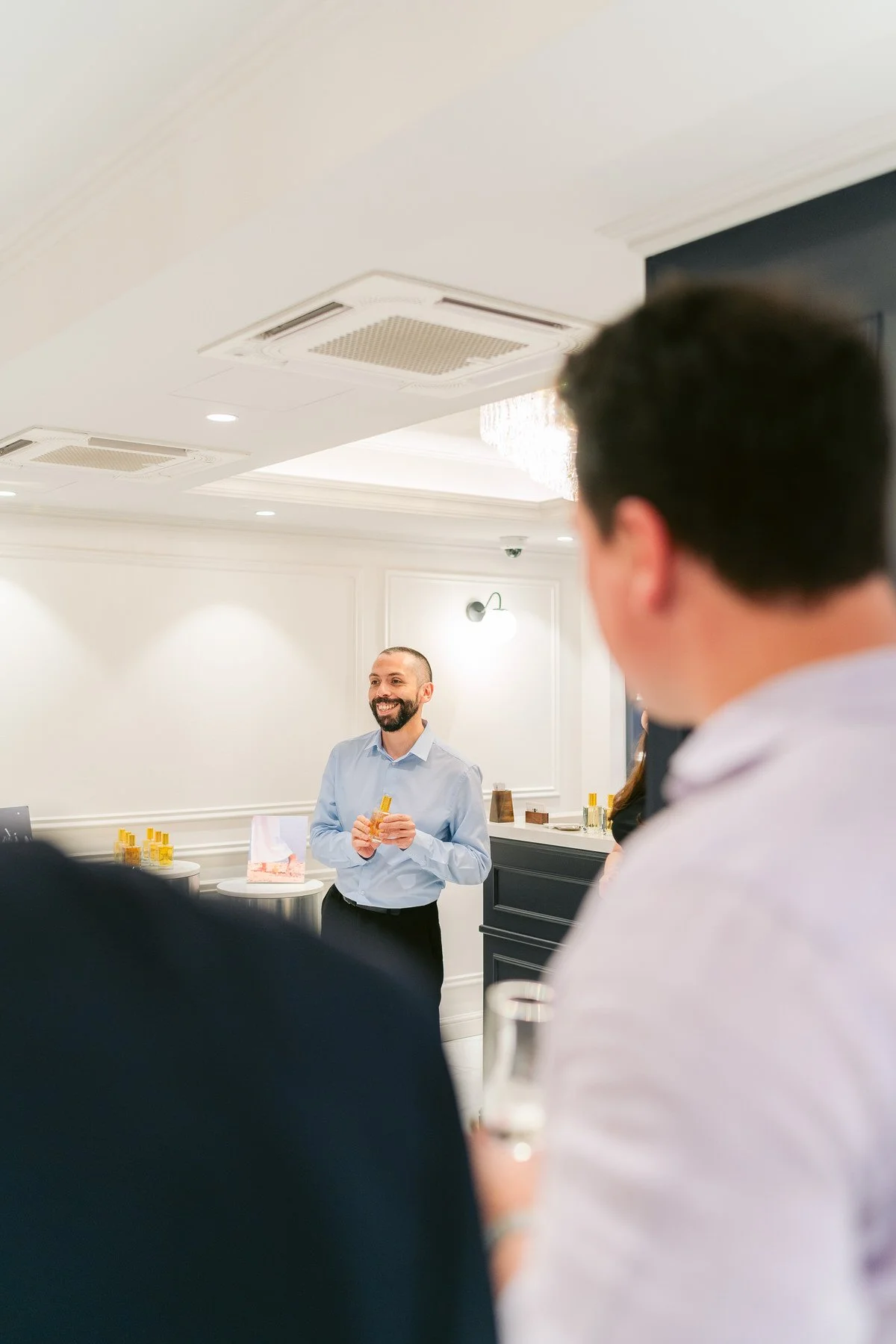 HOMBRE sonriendo y de cabello corto, vestido con camisa azul, de pie en un evento social en interiores, con varias personas a su alrededor, decoraciones blancas y algunos objetos dorados en el fondo.