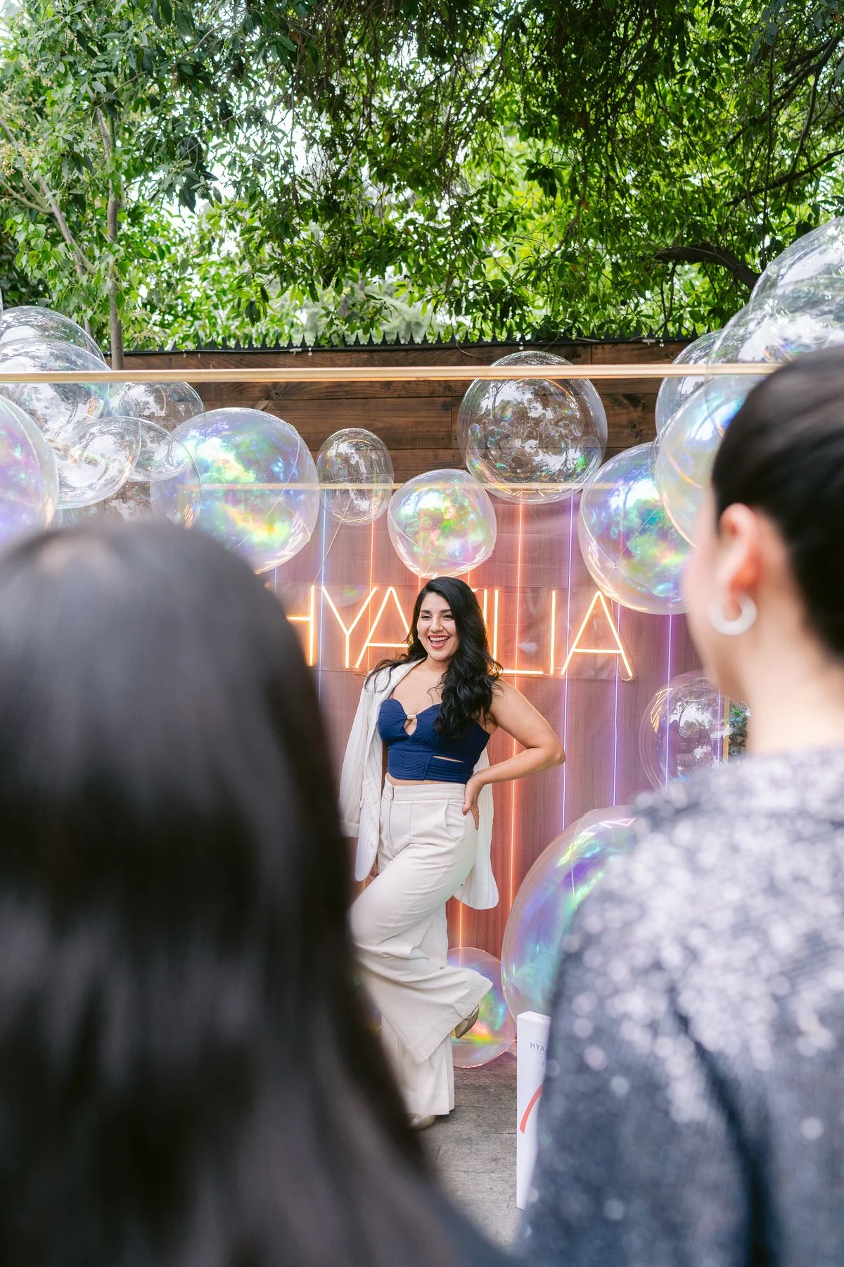 Mujer en un escenario decorado con globos transparentes y un cartel de neón que dice 'YHAMILIA', rodeada de personas en celebración.