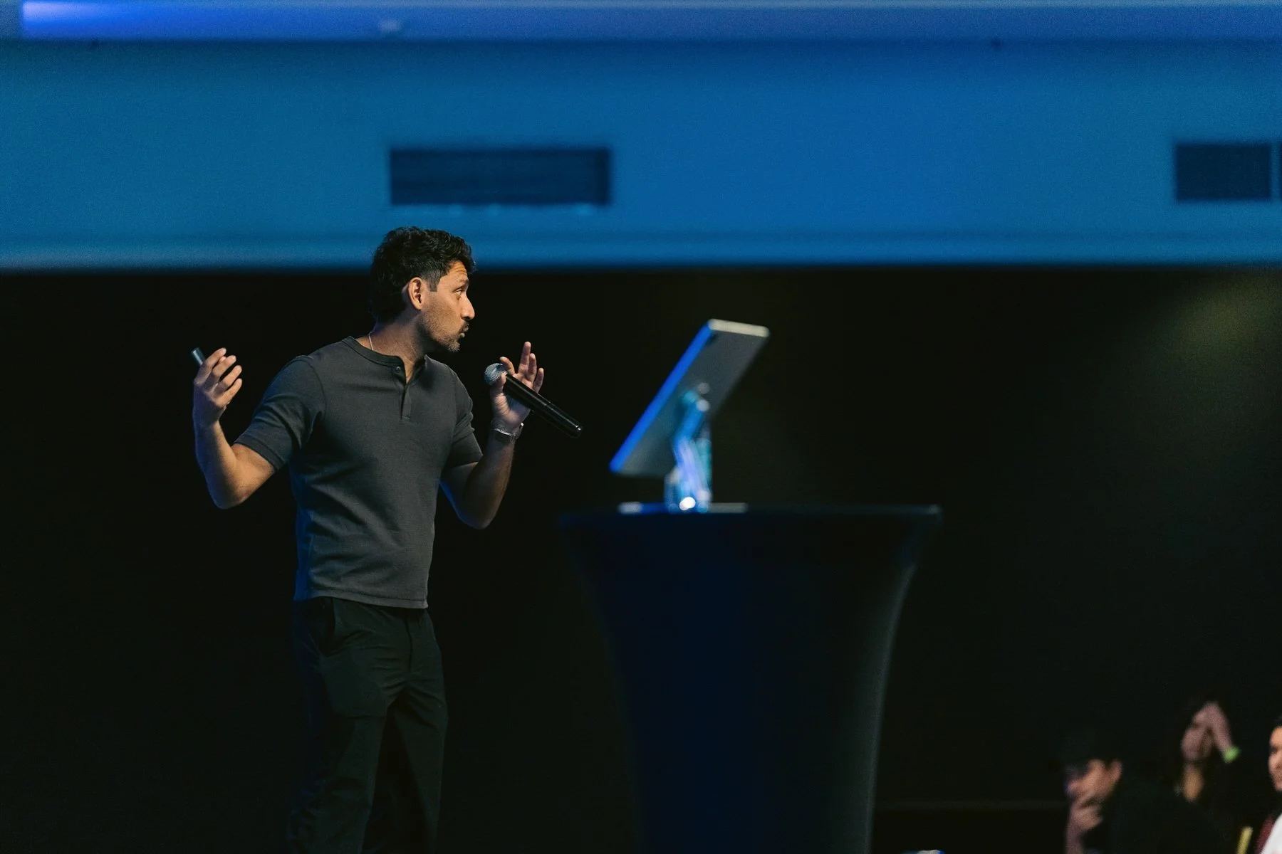 Hombre de pie en un escenario, sosteniendo un micrófono en una mano y usando un control remoto en la otra, frente a una pantalla grande en un ambiente de conferencia o presentación.