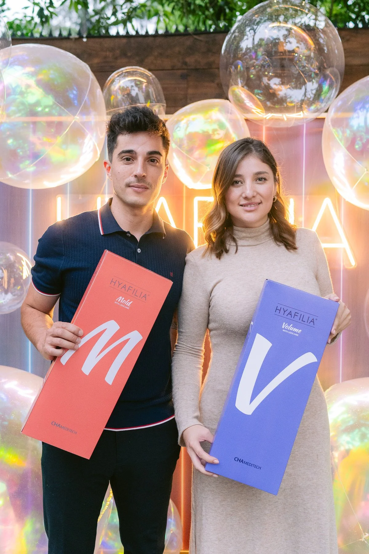 Hombre y mujer sosteniendo cajas de productos Hyafilia frente a fondo con globo de cristal y luces de neón en un evento.