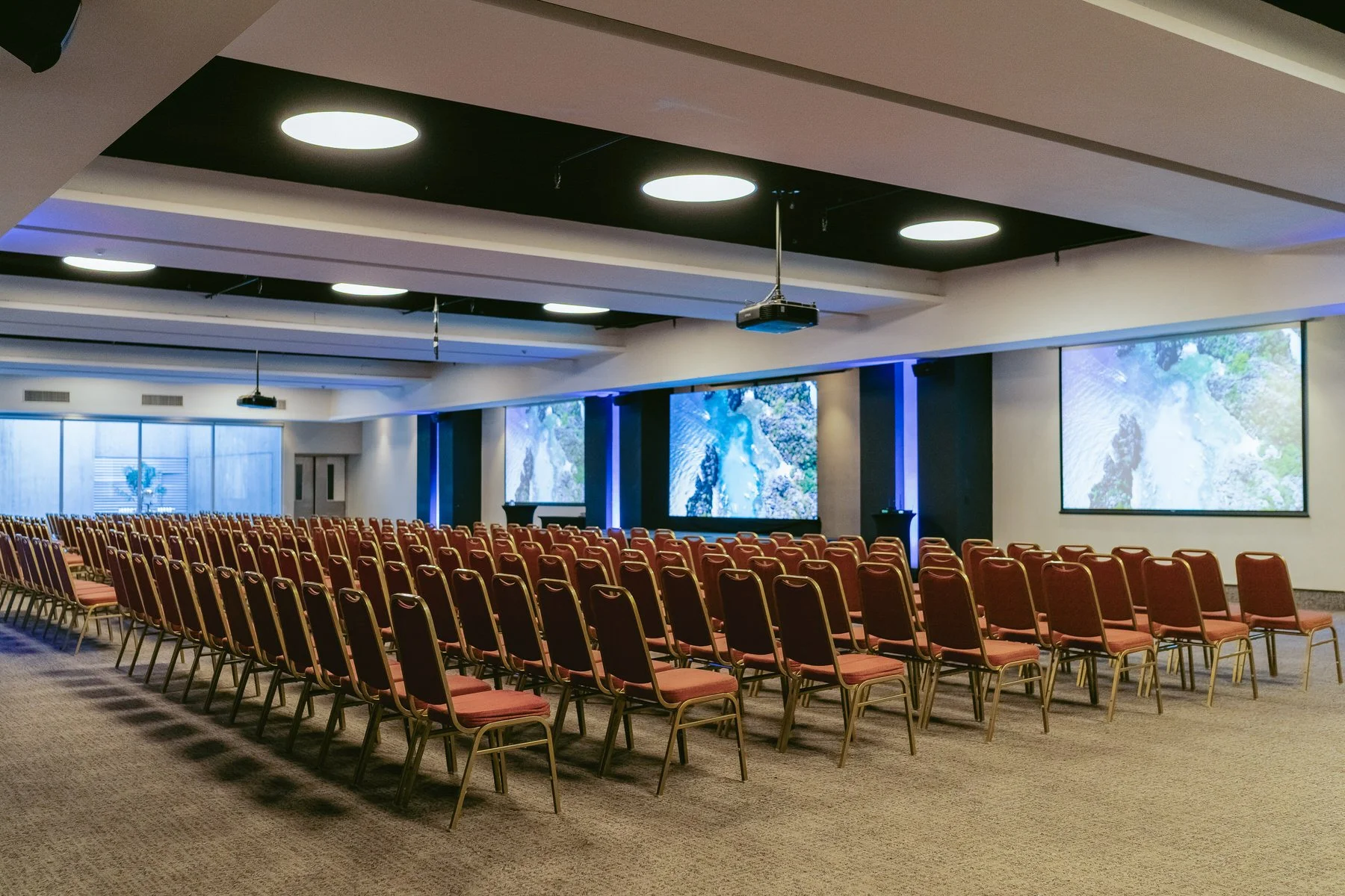 Sala de conferencias con varias filas de sillas y pantallas grandes en la pared.