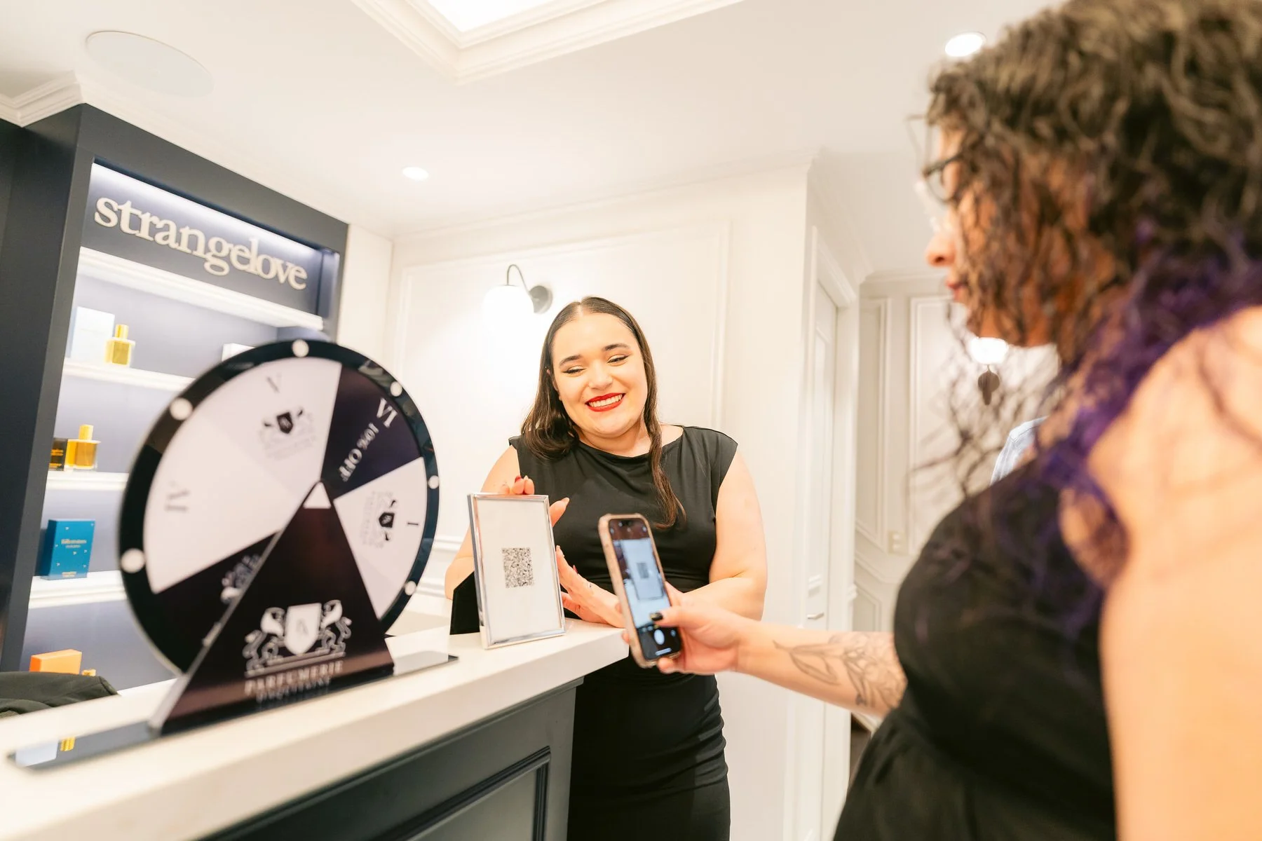 Mujer con vestido negro sonriendo en un mostrador, otras personas tomando fotos, fondo con cartel de 'strangelove' y productos en estantes.