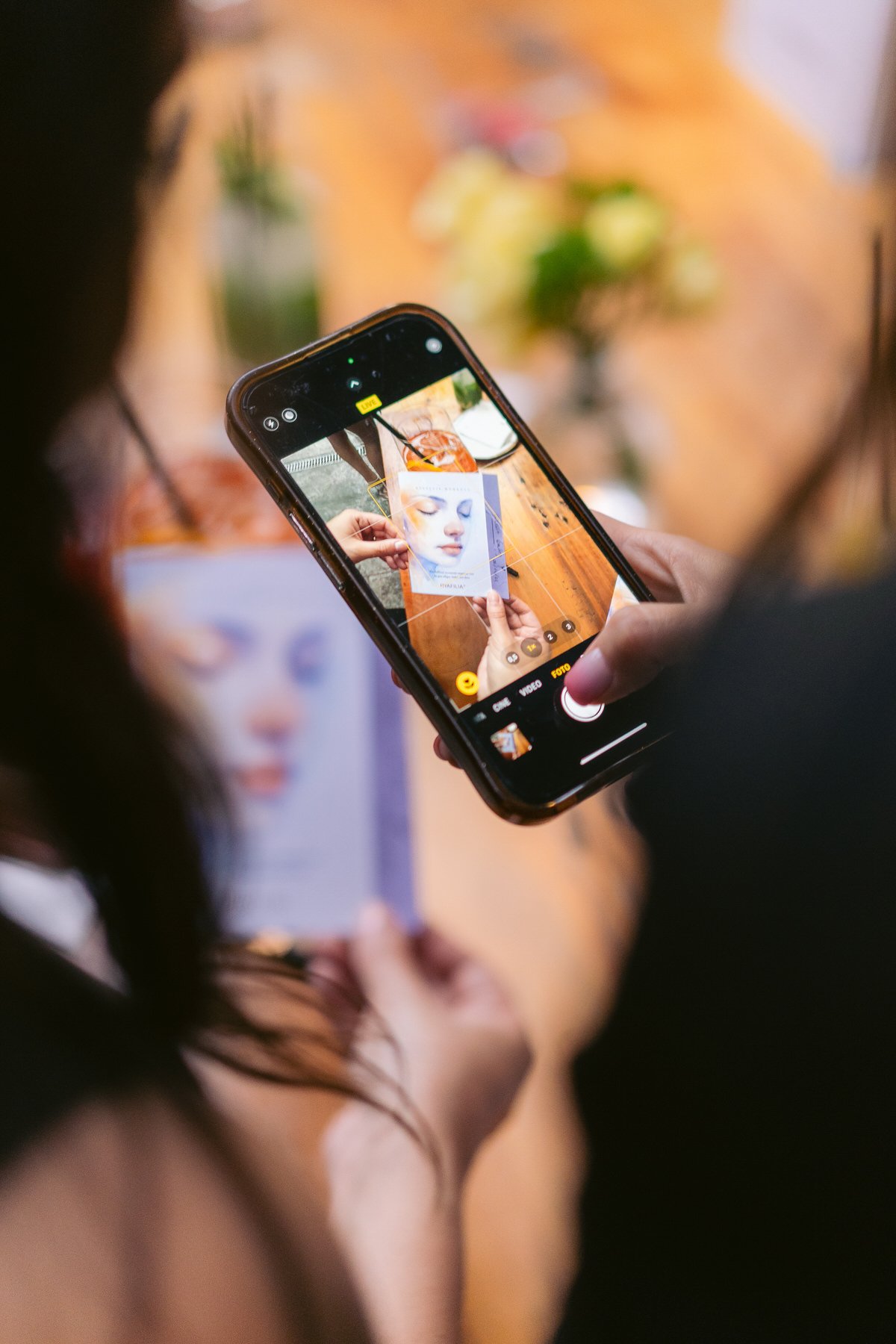 Una persona toma una foto con un teléfono móvil de otra persona que sostiene una ilustración de un rostro. La escena está en una mesa de madera con algunas flores en el fondo.