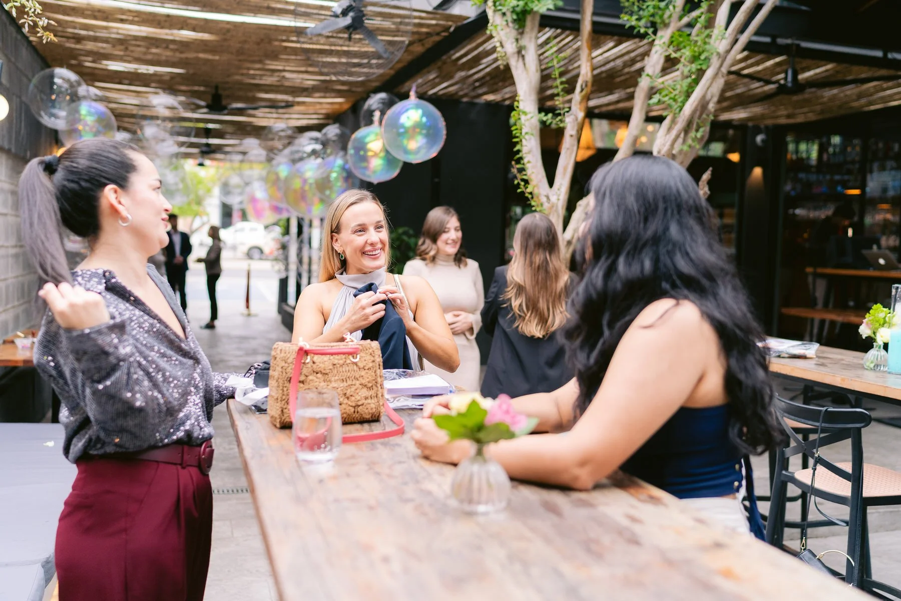 Un grupo de mujeres conversando y sonriendo en un restaurante al aire libre decorado con burbujas y flores.