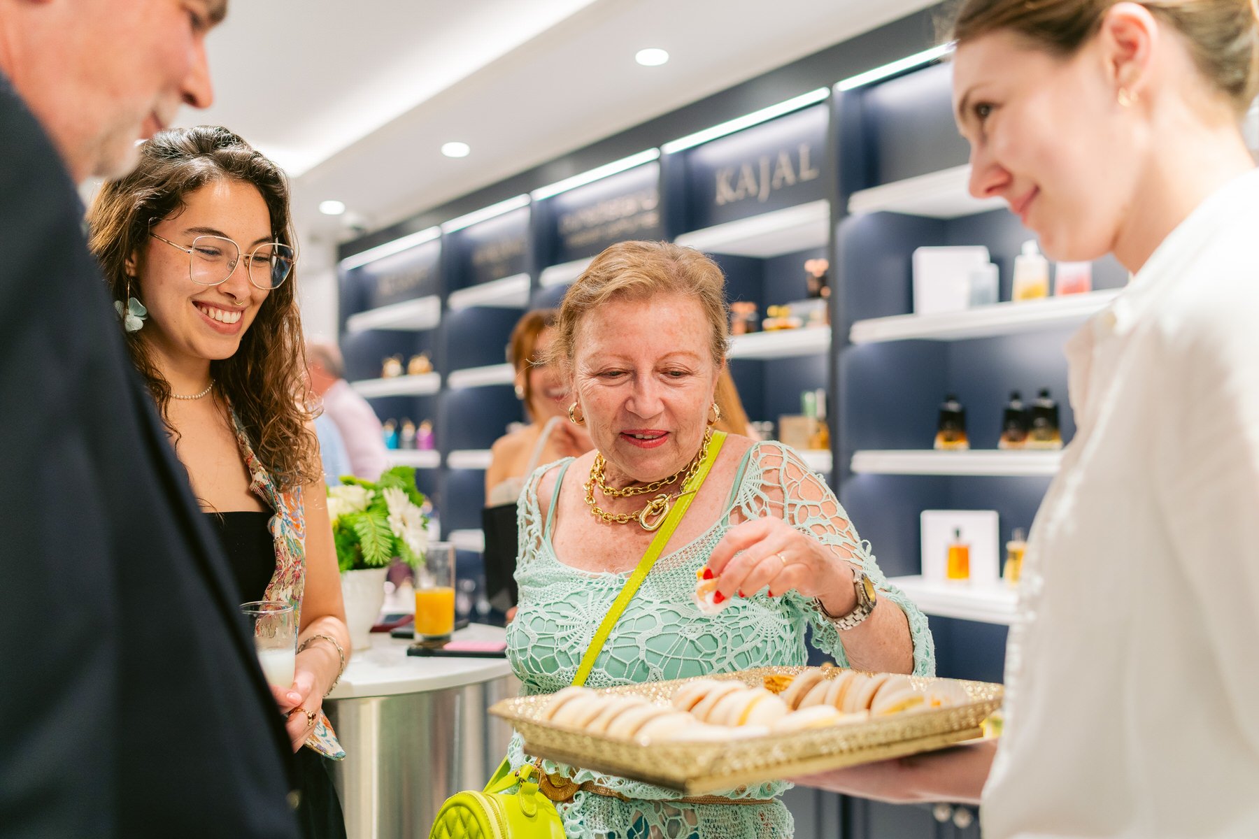 Cuatro personas en una tienda, una mujer de edad media recibe un plato lleno de pequeños alimentos a una joven con gafas y otra mujer con cabello rubio en un centro comercial.