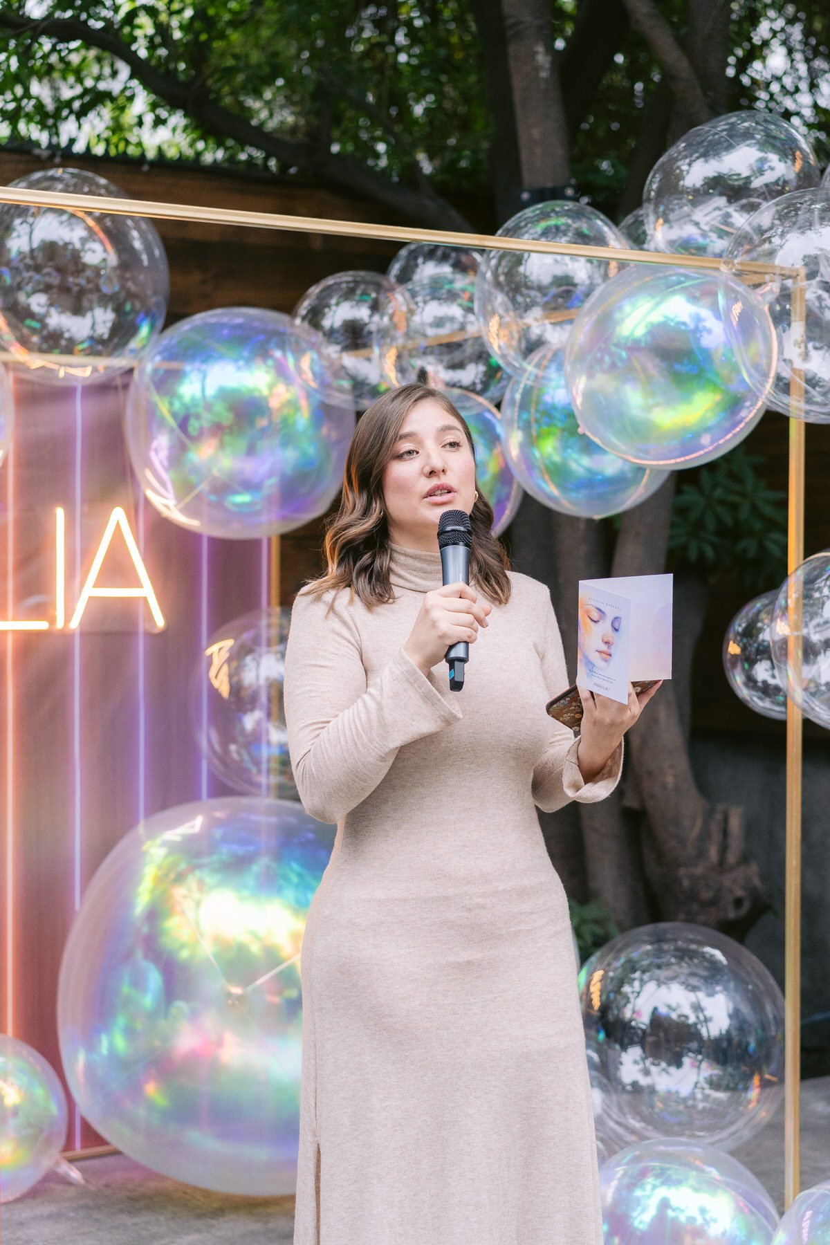 Mujer hablando en un evento con globos de burbujas de colores en el fondo y luces de neón que dicen 'LA'.