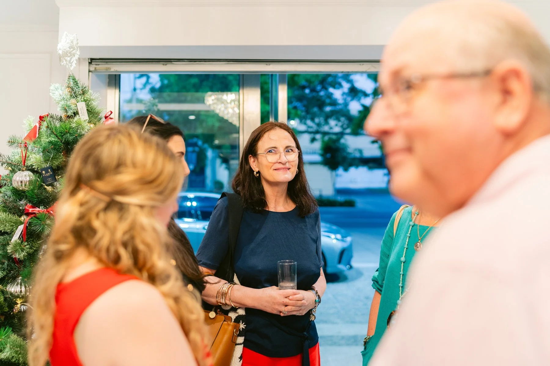 Mujer con gafas y cabello oscuro en una fiesta navideña con árbol de Navidad decorado a la izquierda, otros invitados conversando, y un coche visible afuera a través de las ventanas.