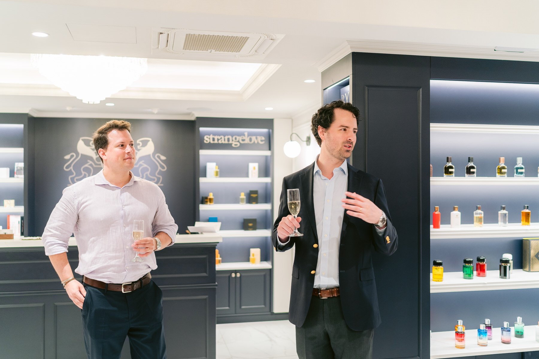 Dos hombres en una tienda de perfumes, uno vestido con chaqueta y el otro con camisa, sosteniendo copas de champagne