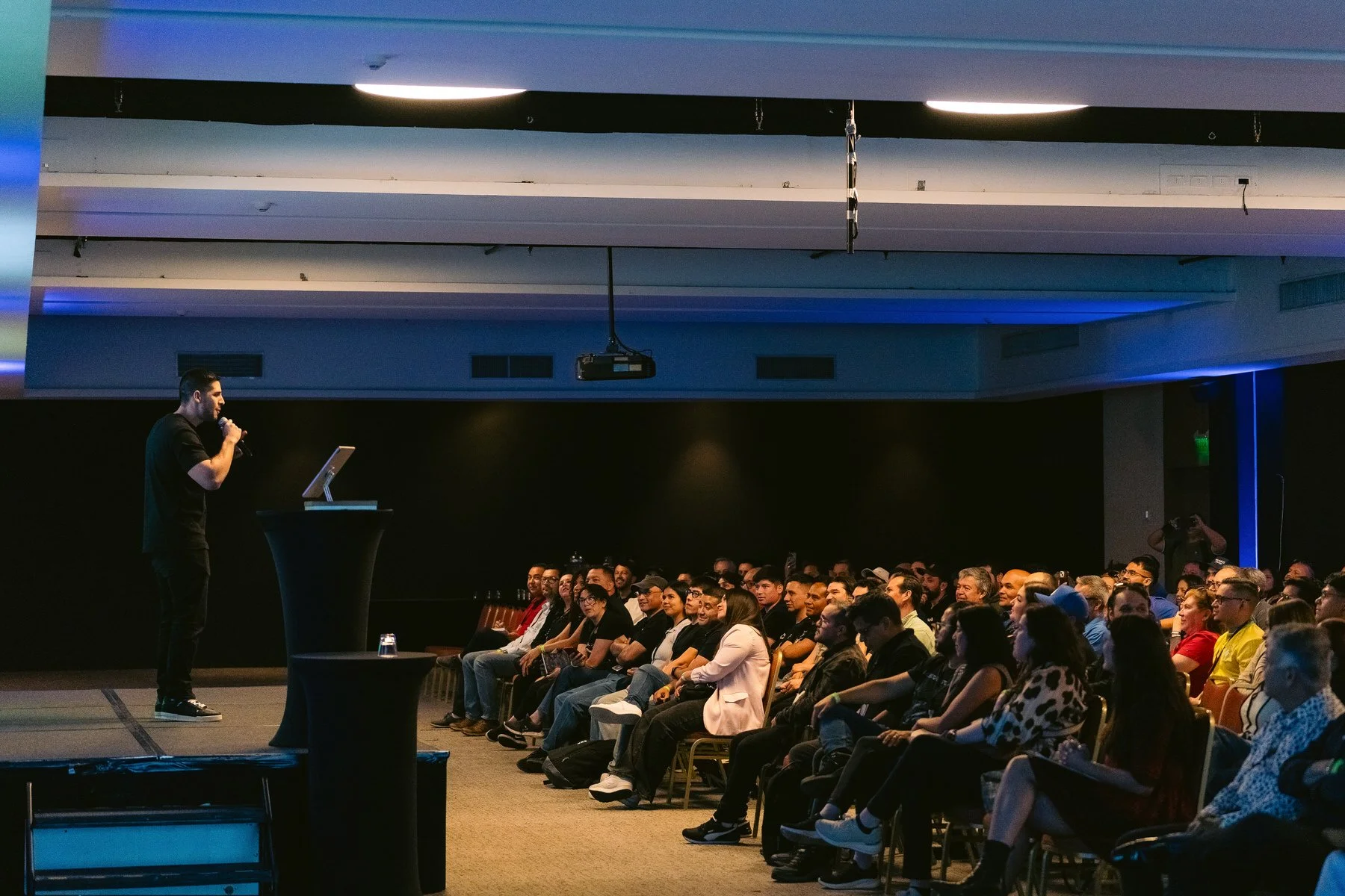 Un conferencista en un escenario presenta frente a una audiencia en un salón de eventos.