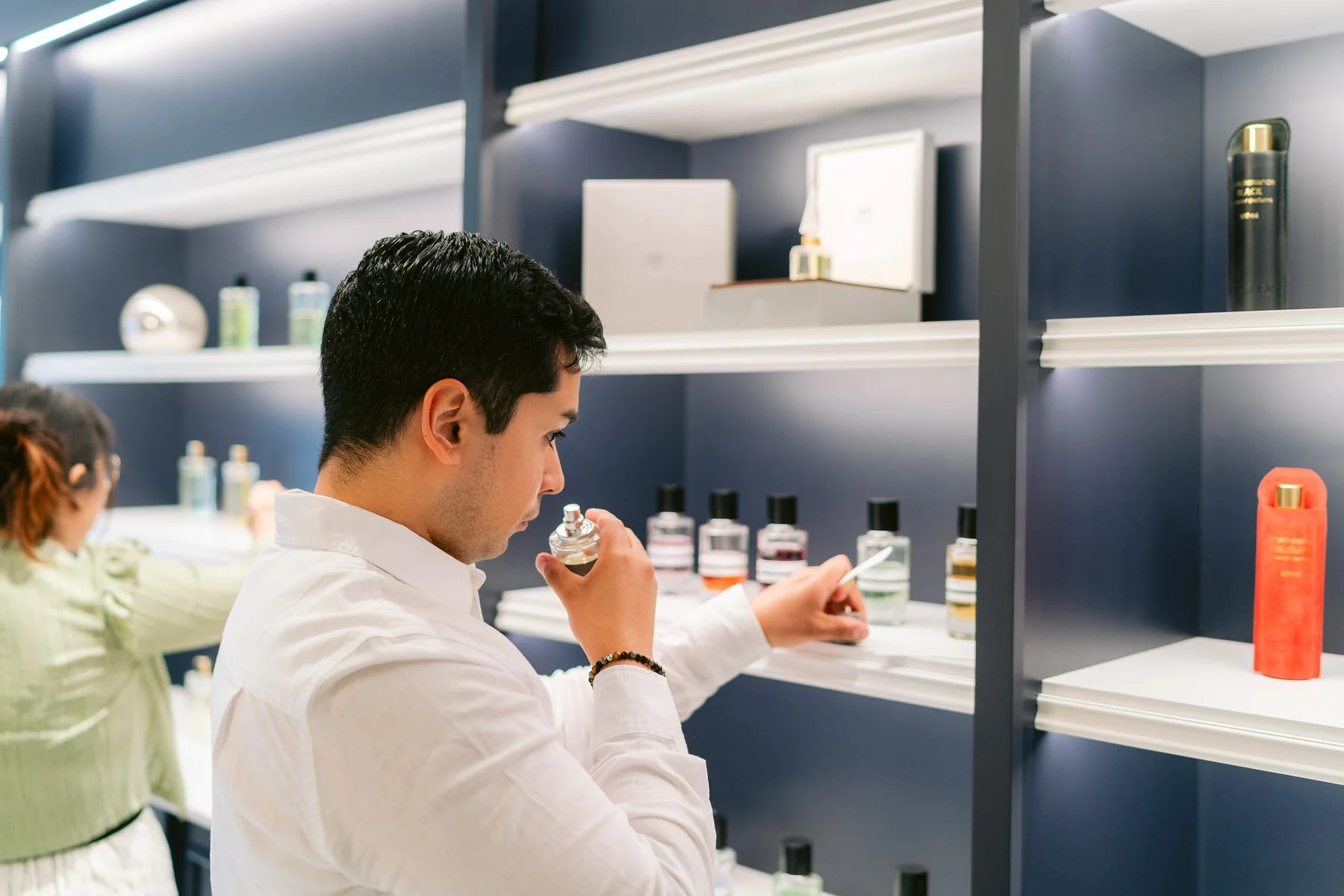 Hombre probándose perfume en una tienda de cosméticos y otros clientes en el fondo.