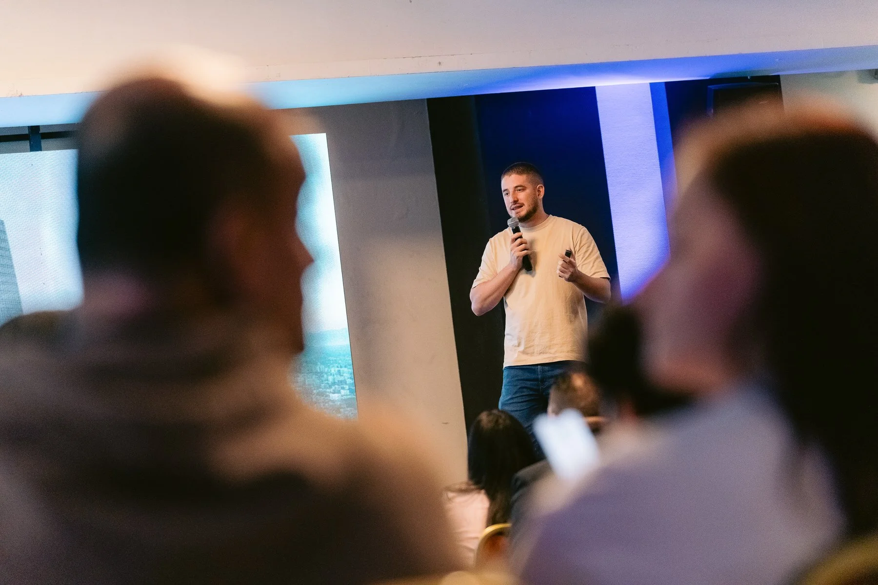 Hombre en camiseta beige dando una conferencia con micrófono en un auditorio con audiencia.