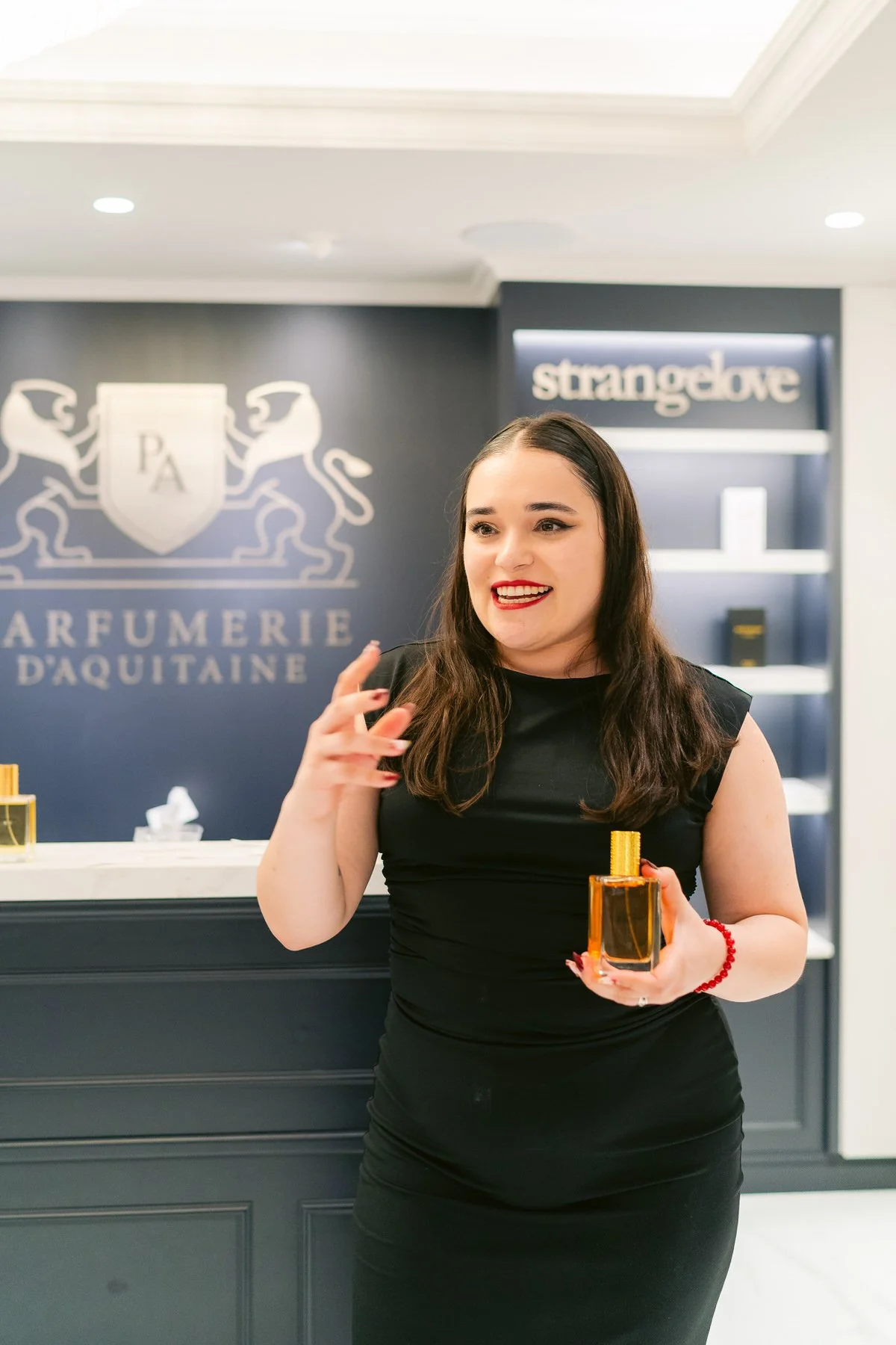 Mujer con cabello largo y oscuro sosteniendo una botella de perfume en una tienda de perfumería, sonriendo y usando vestido negro.