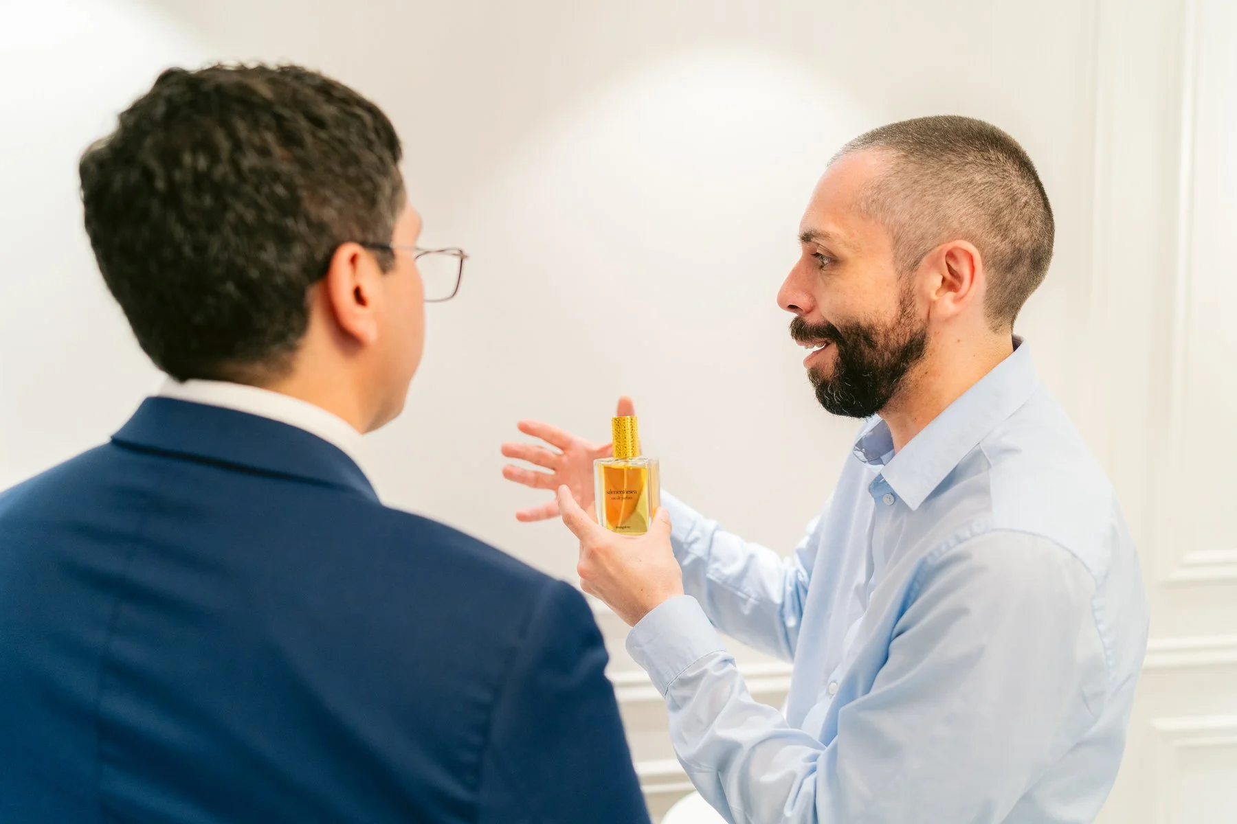 Hombre explicando propiedades de un perfume a otro en una reunión.