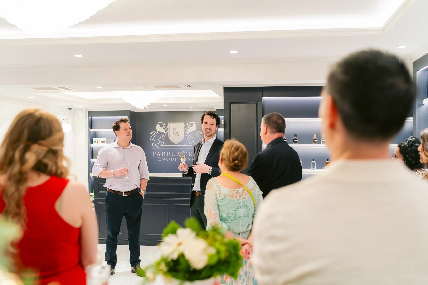 Personas en una reunión social en un espacio elegante, con una pared de fondo que tiene un logotipo con el texto "Parfumerie d'Au...