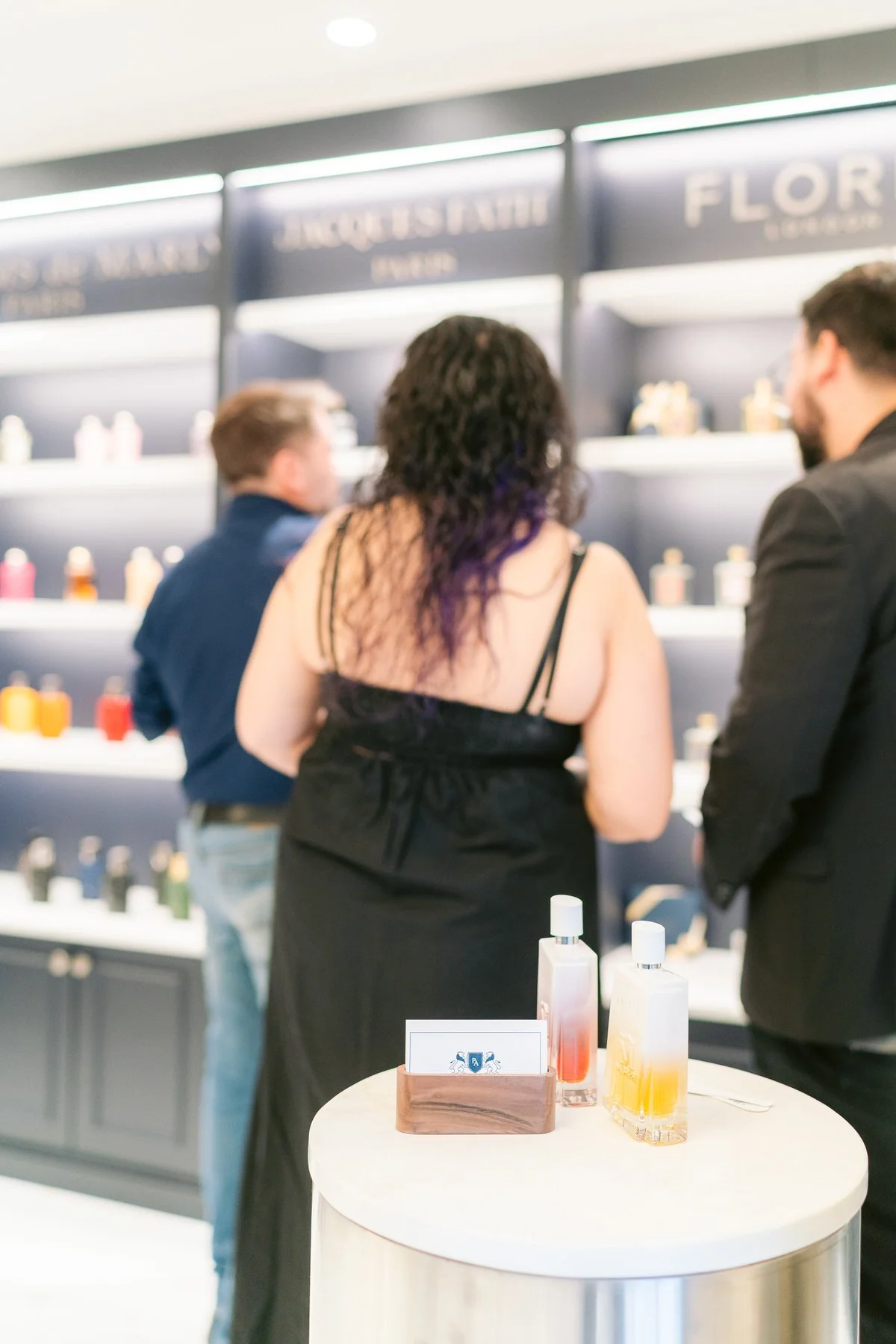 Personas en una tienda de perfumes observando frascos en una estantería, con un mostrador en primer plano que tiene algunos frascos y una tarjeta.