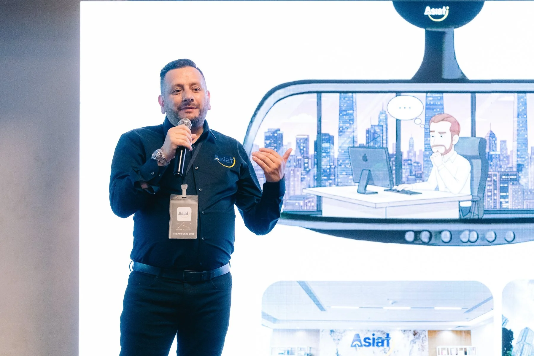 Hombre en una conferencia con micrófono, lleva una camiseta negra con logo de AsiaT. Detrás, una pantalla muestra una caricatura de un hombre en una oficina con vista a la ciudad.