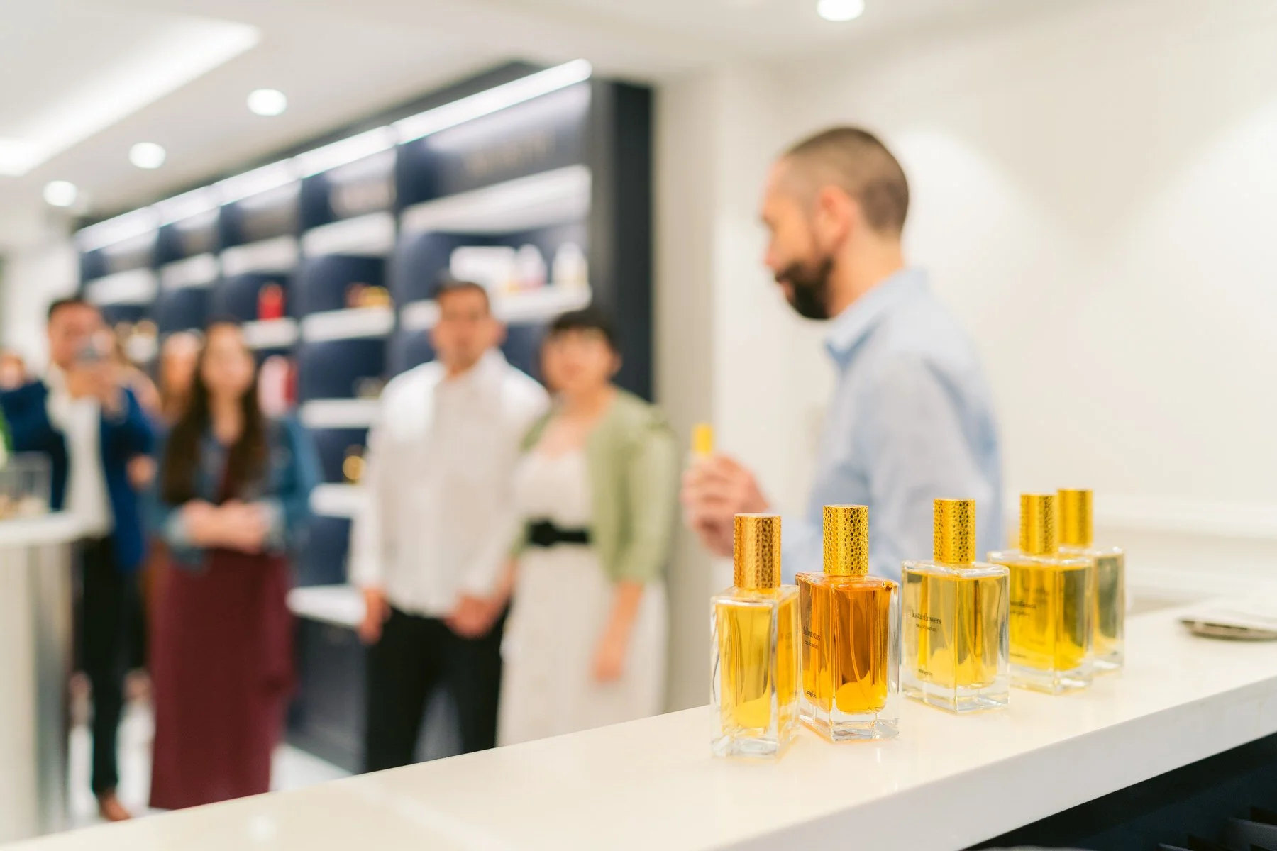 Varios frascos de perfume en primer plano sobre una mesa blanca, con un grupo de personas borrosas en el fondo en un evento o tienda de perfumes.