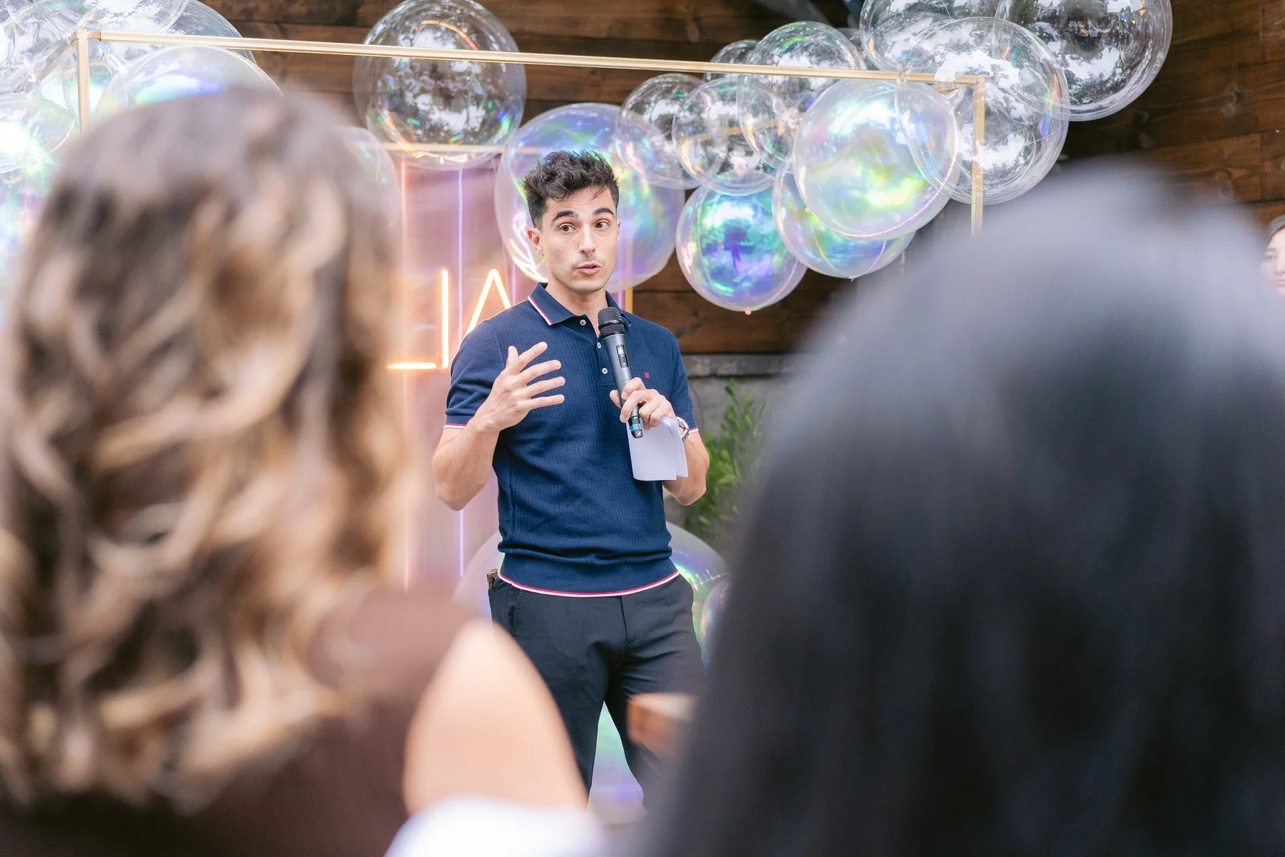 Un hombre joven con cabello oscuro y ropa azul hablando en un micrófono frente a un grupo, decorado con globos transparentes y luces en un evento indoor.