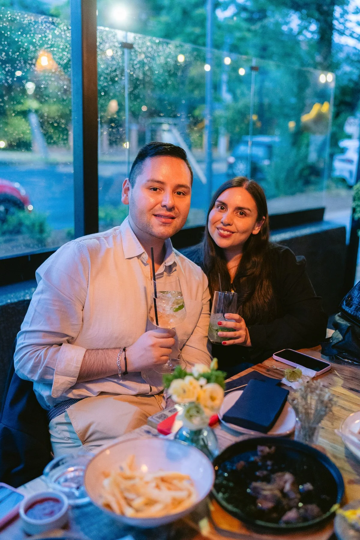 Una pareja sentada en un restaurante con mucha luz azul, con comida y bebidas en la mesa, y un fondo con ventanas y vistas exteriores.