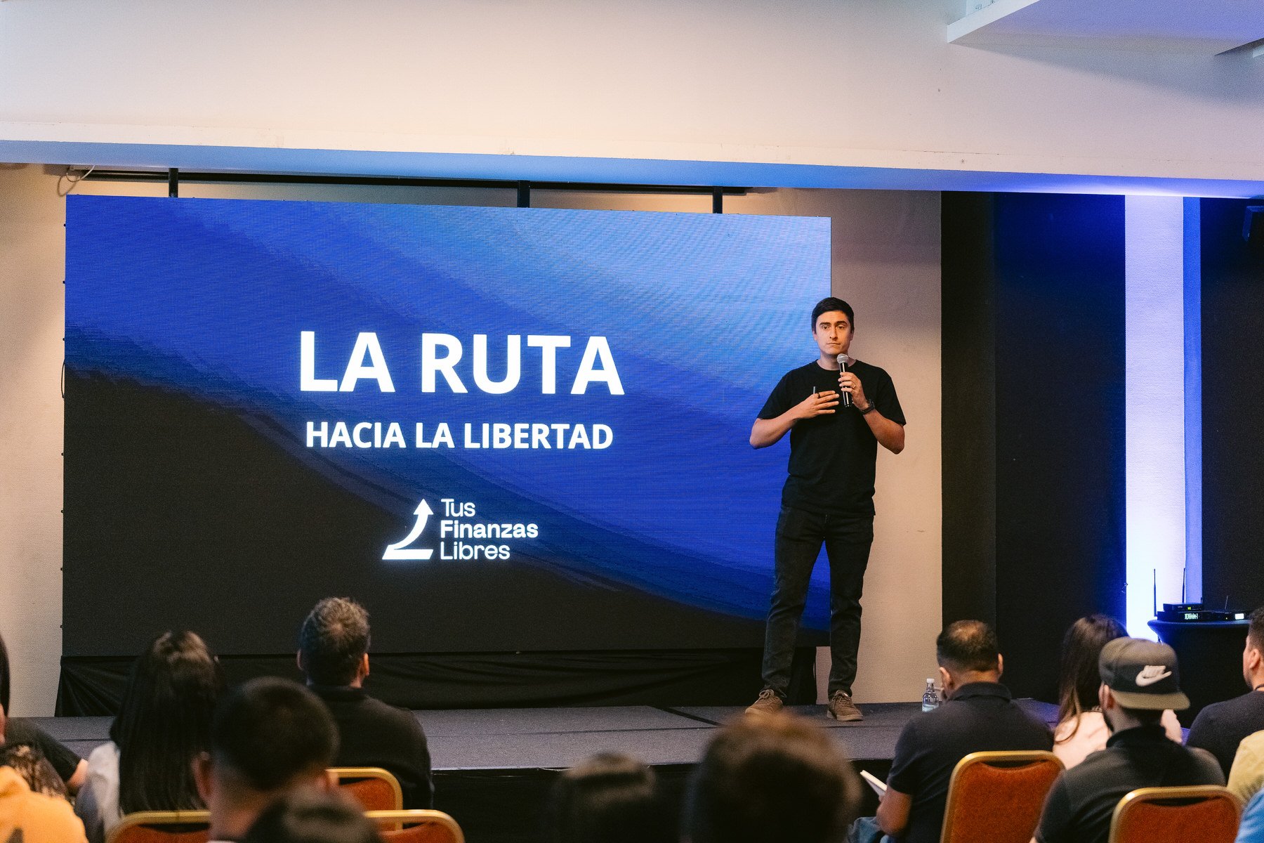 Hombre en escenario dando una conferencia frente a una pantalla gigante que dice 'LA RUTA HACIA LA LIBERTAD' y un logo de 'Tus Finanzas Libres'.