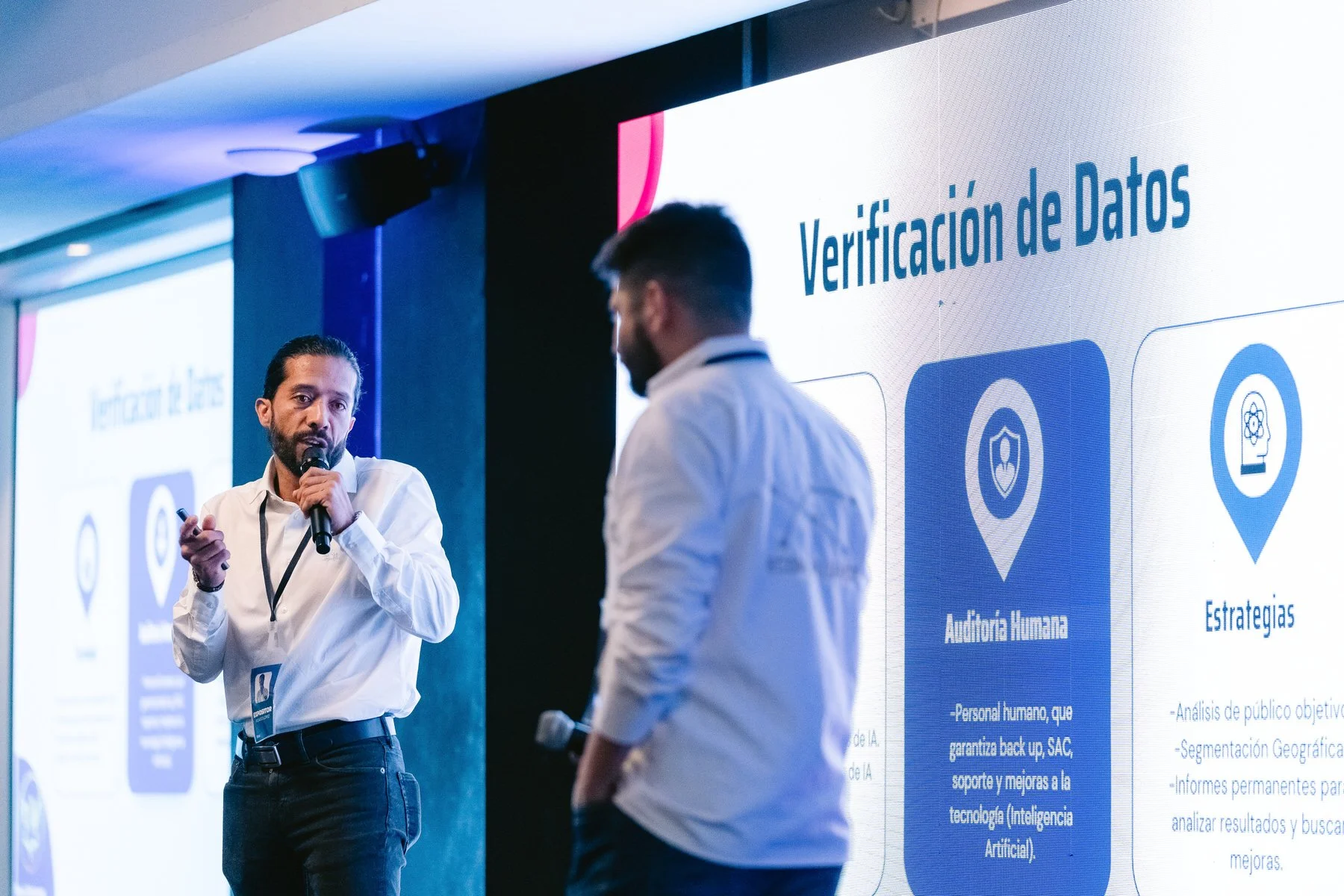 Dos hombres en una conferencia de negocios, uno hablando con micrófono y el otro aparentemente escuchando, con pantallas de presentación en el fondo que muestran información sobre verificación de datos y audiencia humana.