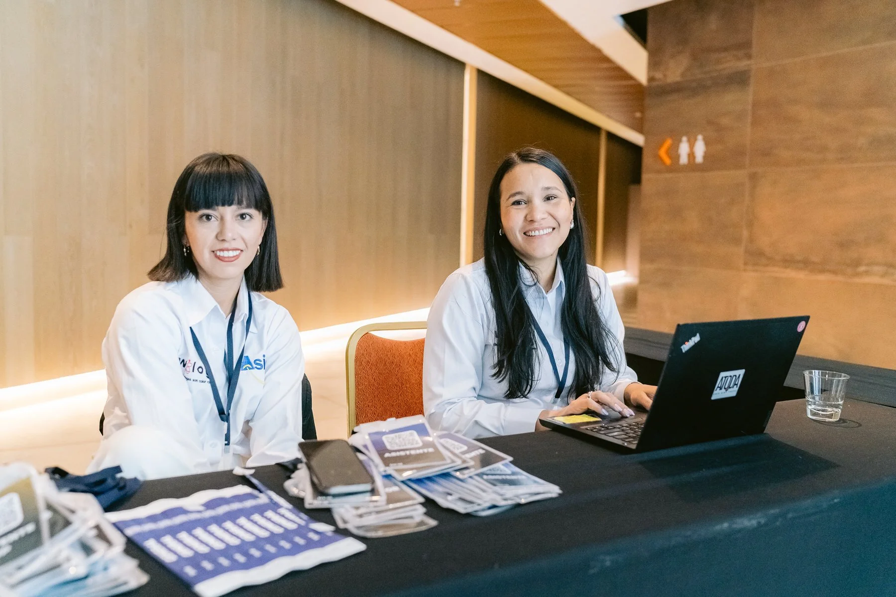 Dos mujeres sentadas en una mesa, una con una computadora portátil y la otra con folletos y teléfonos, en un entorno interior con paredes de madera y señalización de baños en el fondo.