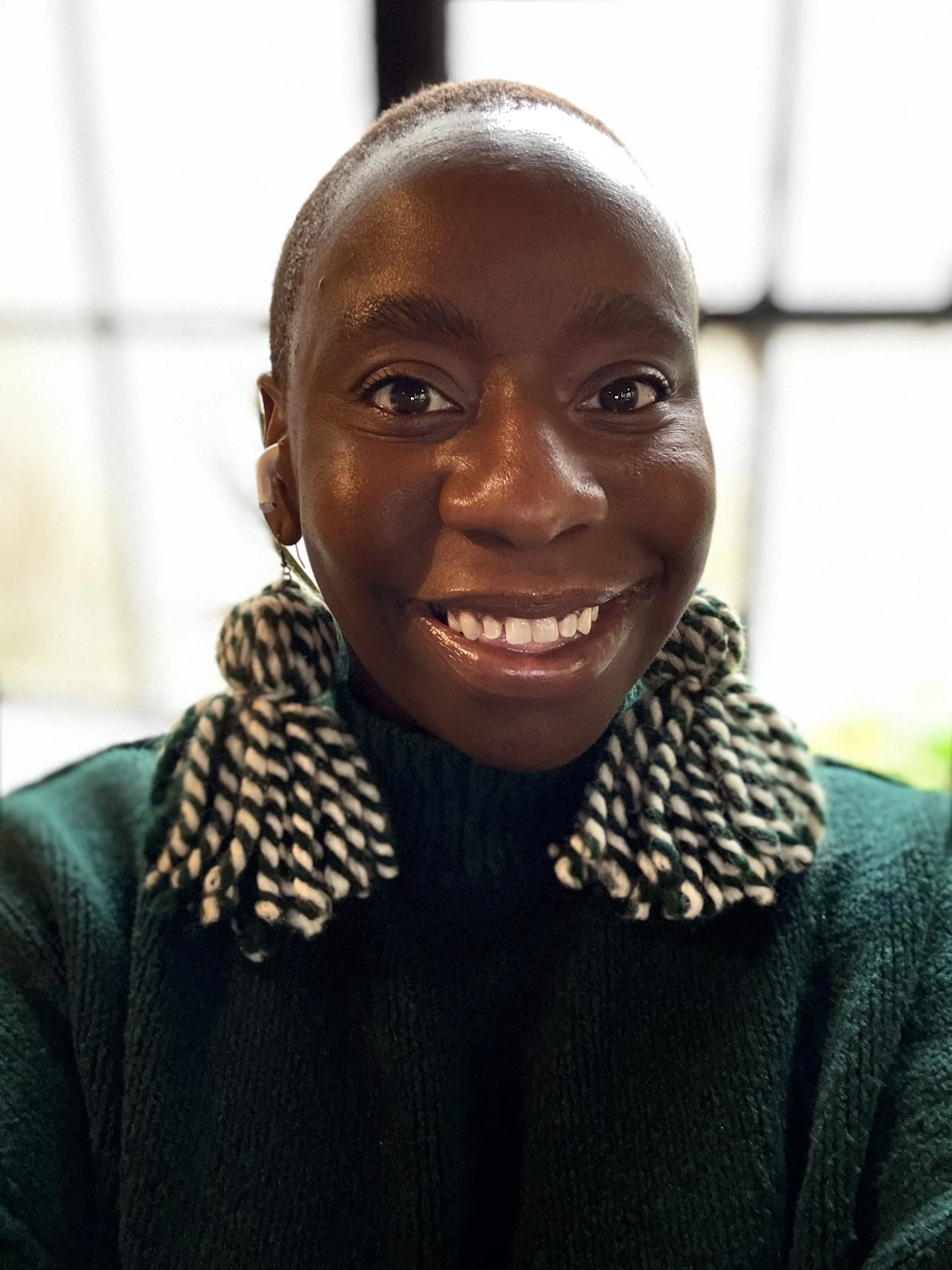 the image shows a smiling Black woman wearing a green sweater with yarn pompom earrings