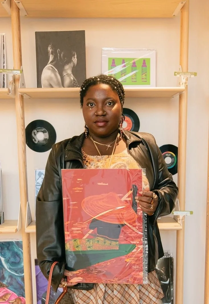 Woman holding a colorful artwork of a boat on water, standing in front of wooden shelves with framed art and records.