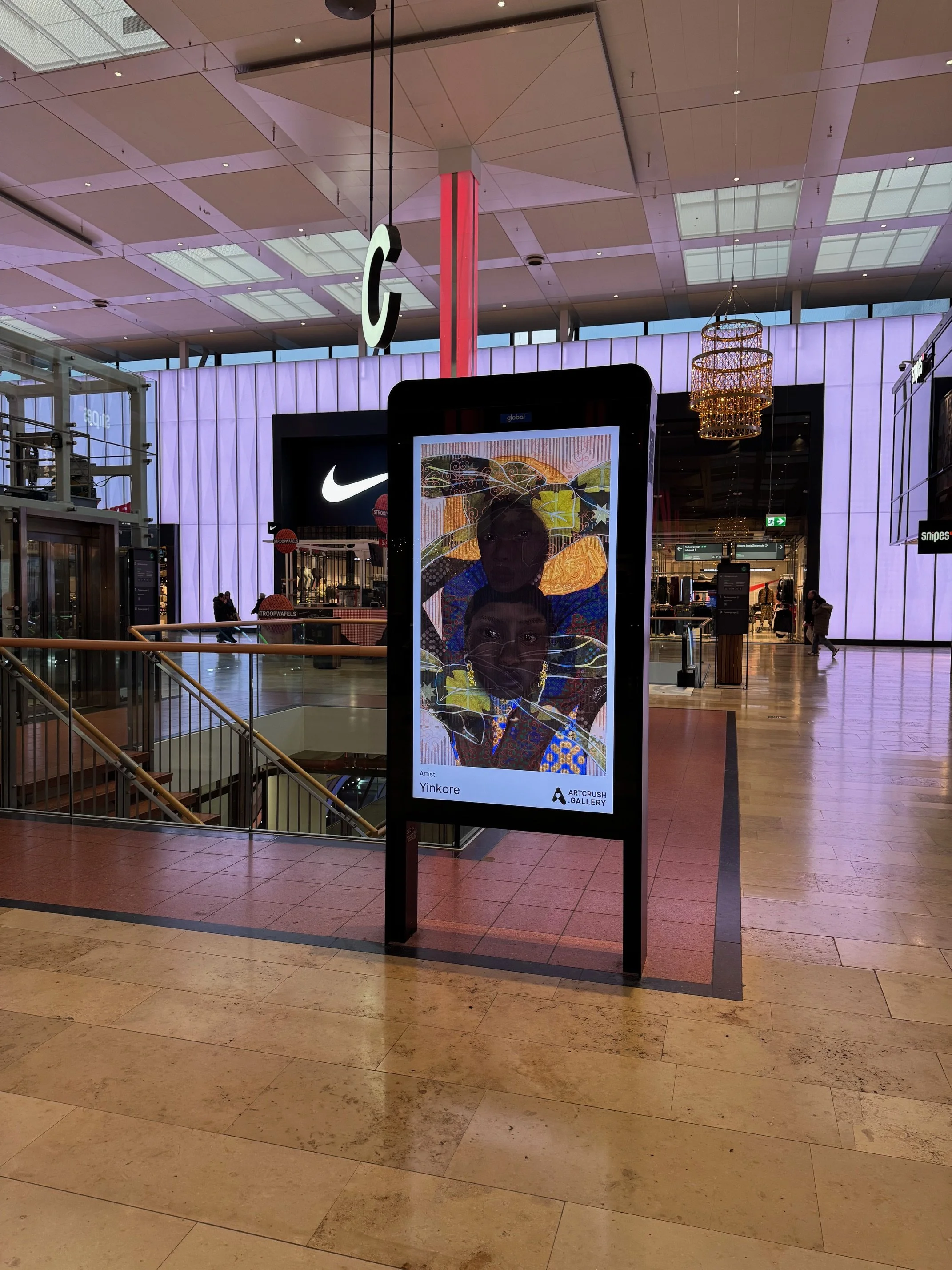 Digital billboard displaying artwork of two faces, one with a dark complexion and one with a lighter complexion, with colorful abstract background. The billboard is in a shopping mall near a Nike store.