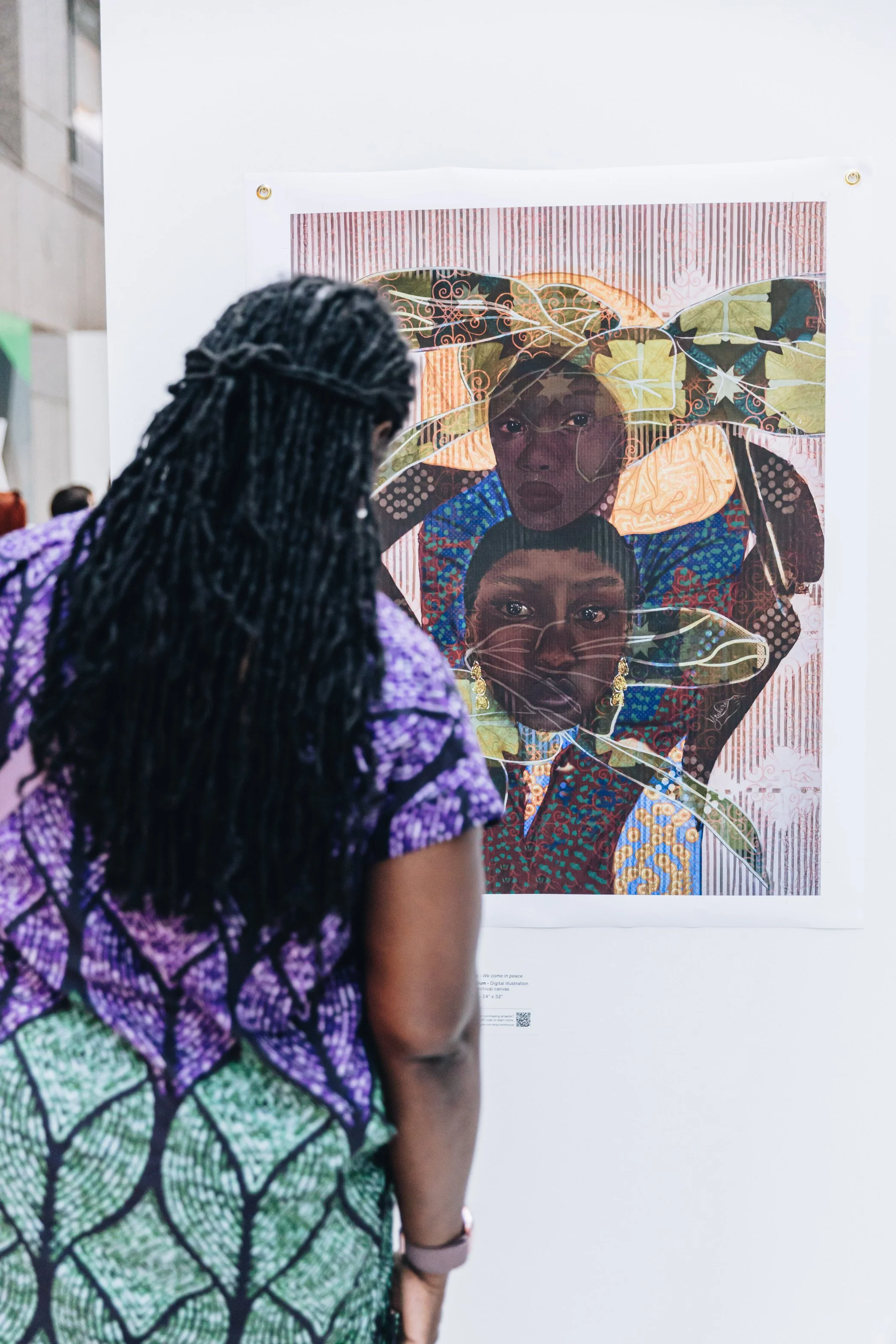 A woman with braided hair wearing a purple and green patterned outfit looks at a colorful digital illustration of two women with large leaves on their heads, displayed on a white wall.